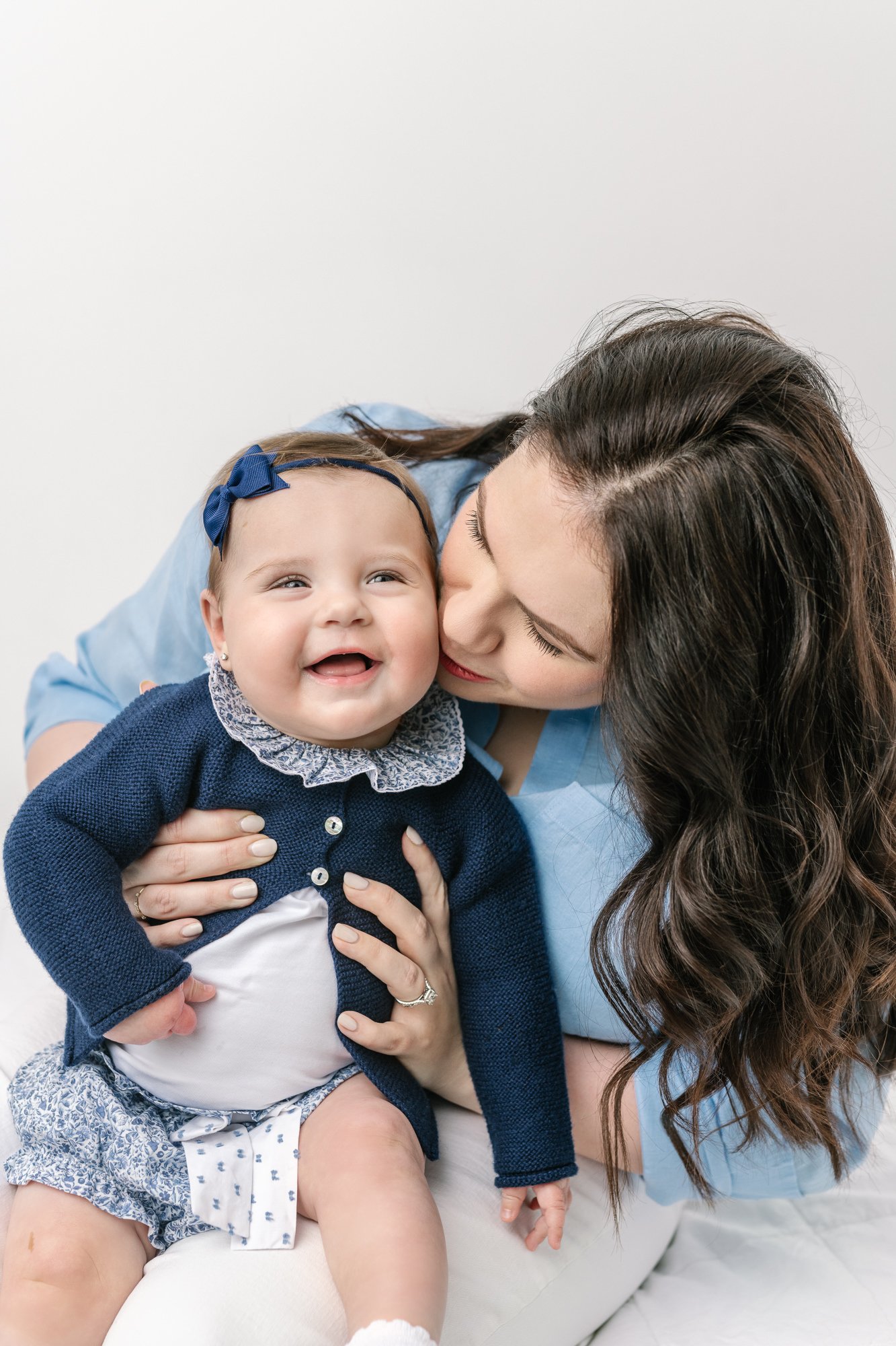  Cutest baby girl sits on her moms lap with a huge smile on her face during Nicole Hawkins Photography session in New Jersey. Mom leans in to kiss daughter’s face. Both are wearing blue. #babystudioportraits #studionewborns #NewJerseyPhotographers #N