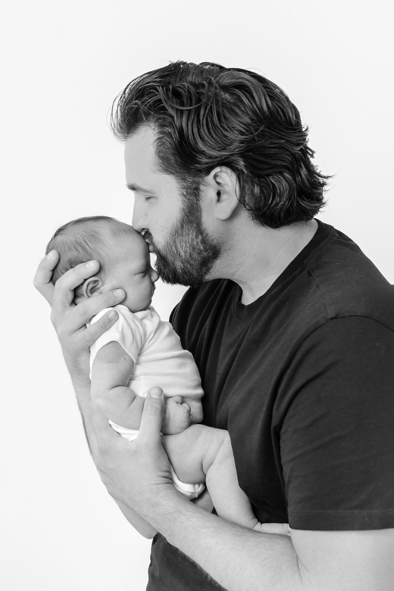  A dad cradles his newborn while giving the baby a kiss on their forehead. Dad and baby connect in a loving, timeless moment during newborn studio session by Nicole Hawkins Photography. Photography session in New Jersey. #newbornportraits #newbornstu