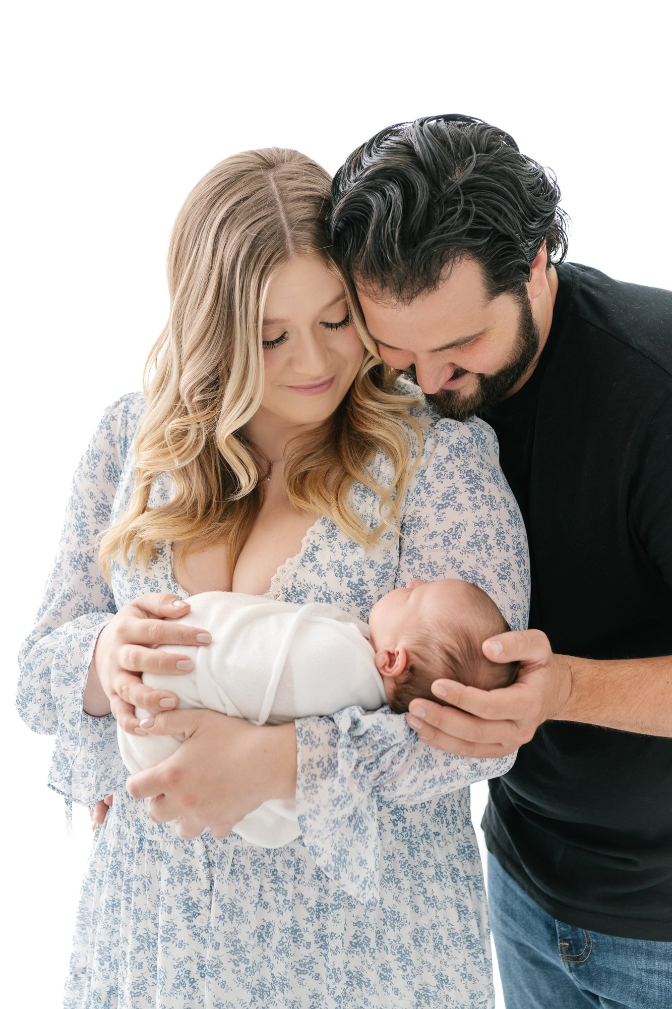  Parents stand shoulder to shoulder while mom holds young baby in this timeless portrait. Dad has a  smile on his face while looking at newborn baby in New Jersey studio session shot by Nicole Hawkins Photography. #newbornportraits #newbornstudioshoo