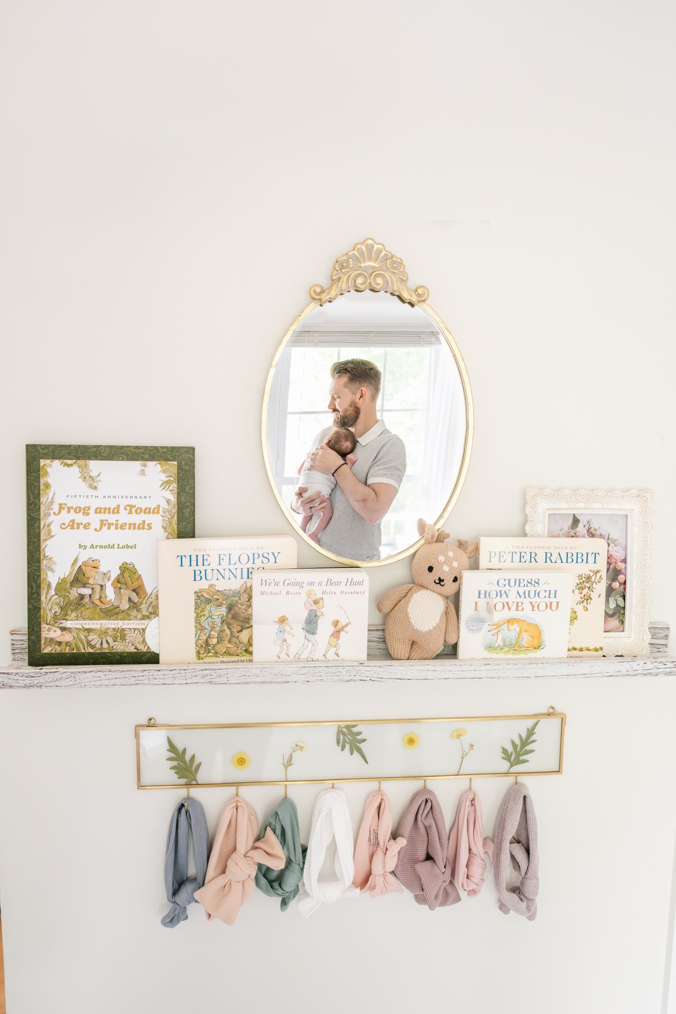  The cutest wall in the new newborn nursery. Shelf with children’s books. Floral shelf with hooks that displays all sorts of darling, classic bows. Oval gold mirror shows reflection of sweet dad holding newborn daughter. Captured by Nicole Hawkins ph