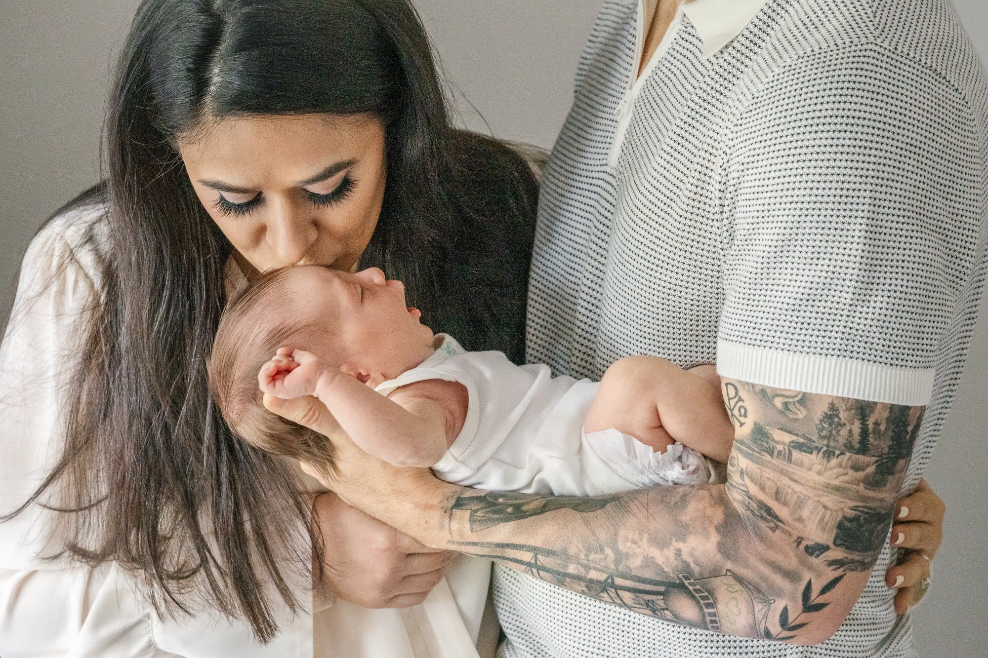  Family dressed in neutrals for in-home newborn photo session with Nicole Hawkins photography. Mom tenderly places a kiss on newborn daughter’s head. Photo showcases dad’s sleeve of intricate tattoos. #centralNewJerseyphotographer #inhomeportraits #n