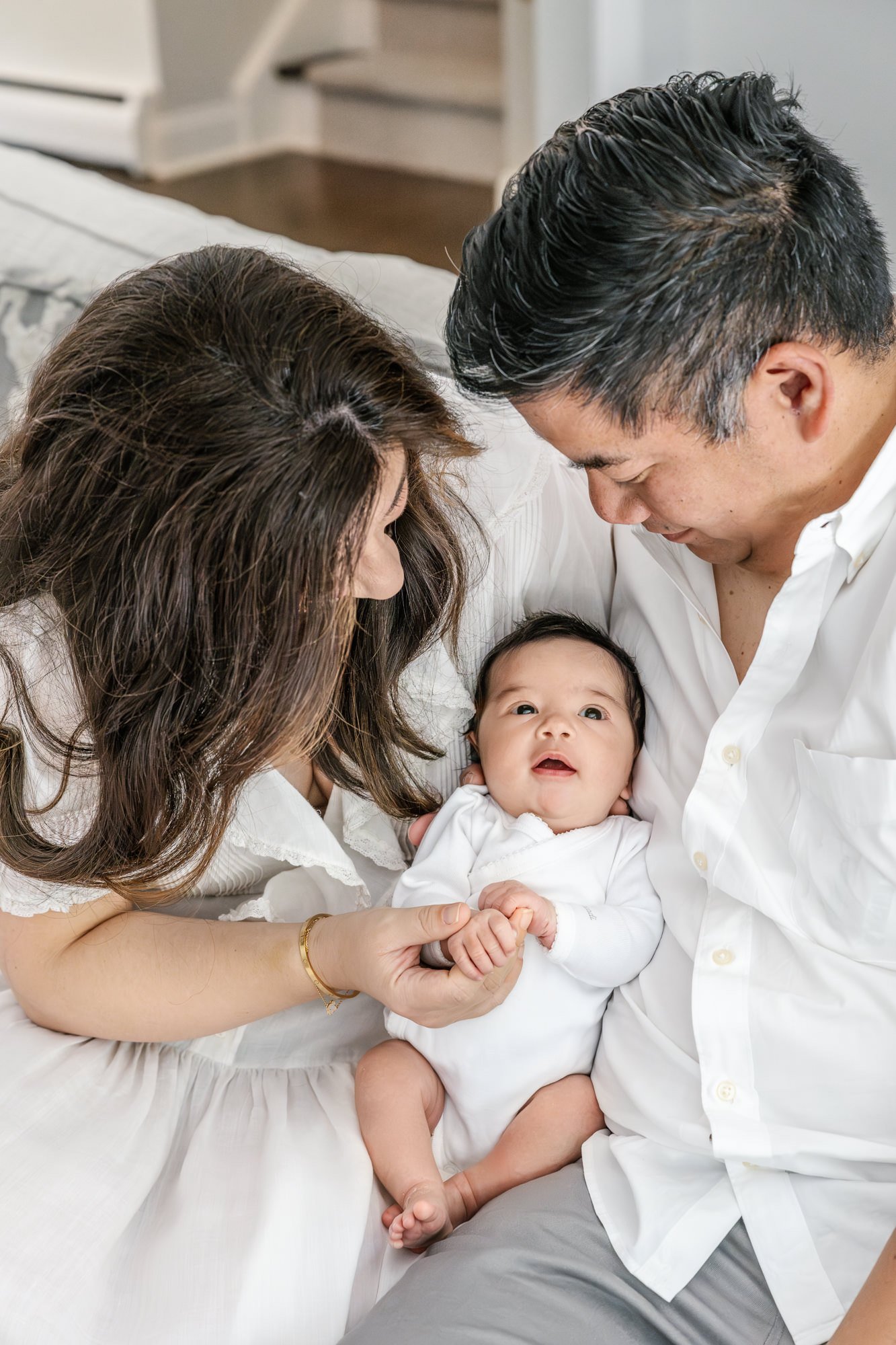  Mom and Dad sit on the bed in their master bedroom while gazing lovingly at their beautiful newborn daughter. #newyorkcityphotographer #familyportraits #newbornportraitsession #inhomephotography #familyoffive #familyposeinspo #newjerseyfamilyphotogr