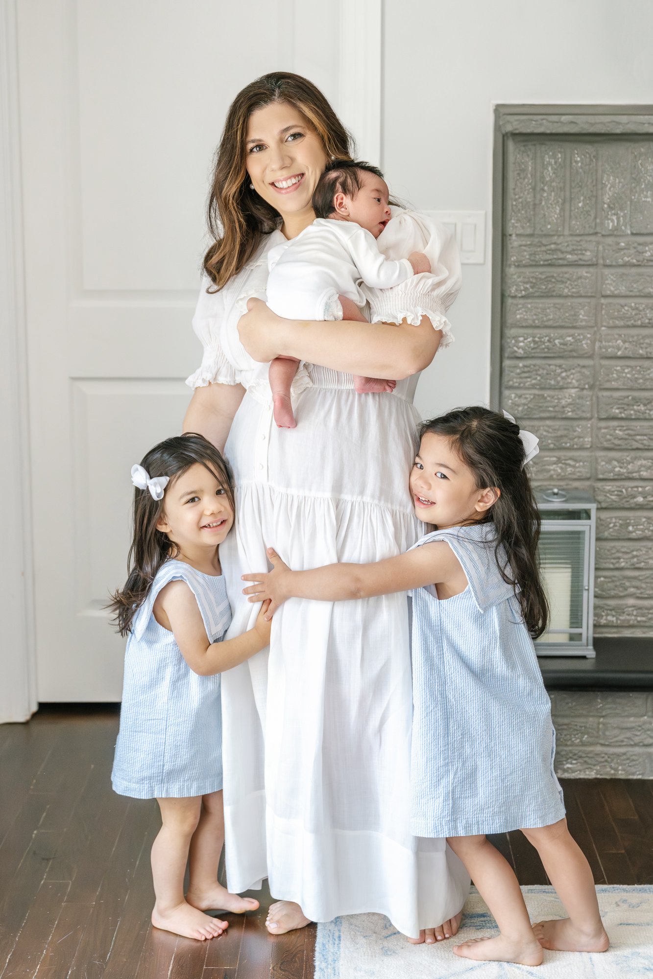 Beautiful mom stands holding her newborn daughter in a white onesie while her two young daughters give her big hugs around her legs. #newyorkcityphotographer #familyportraits #newbornportraitsession #inhomephotography #familyoffive #familyposeinspo 
