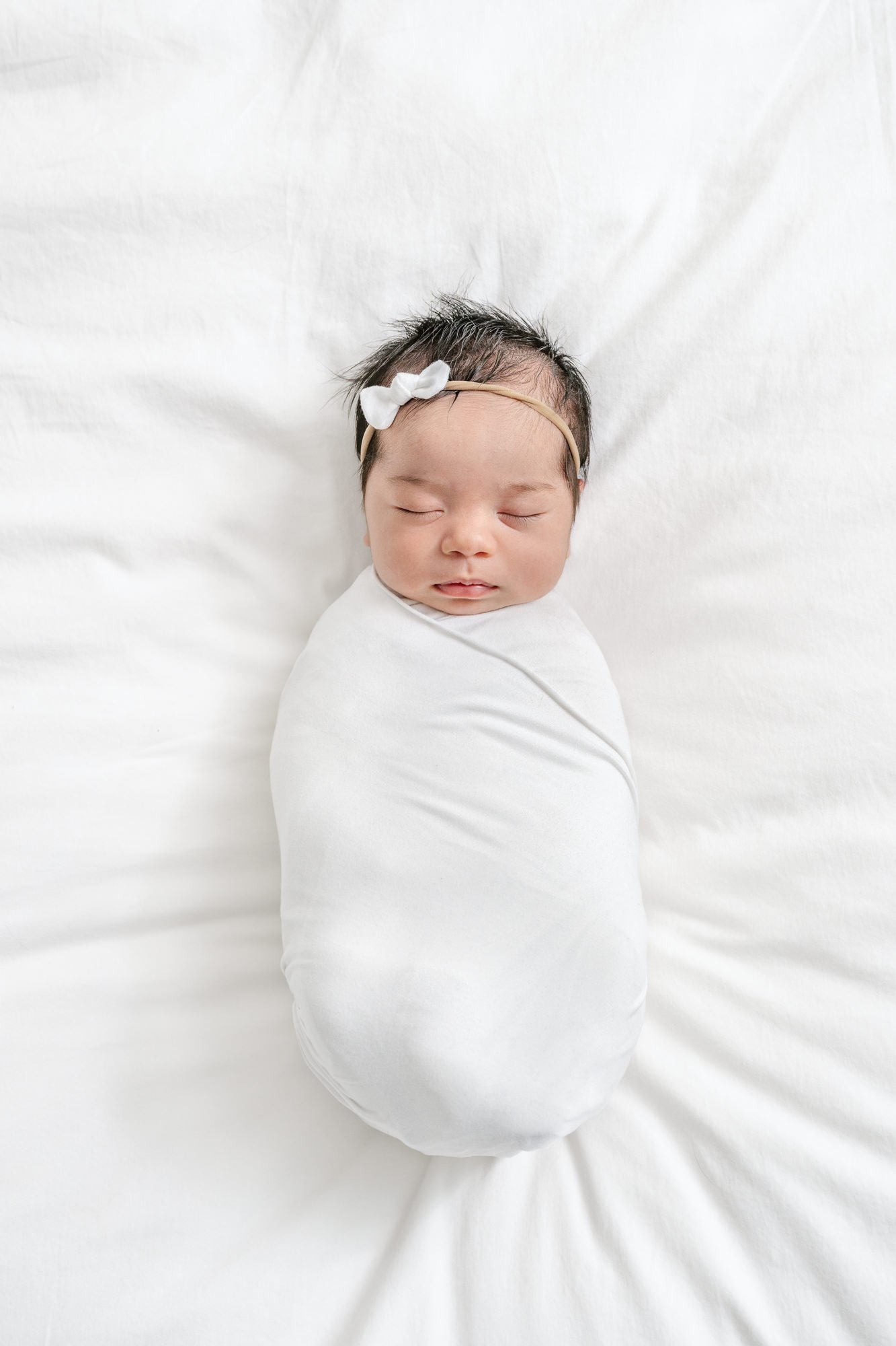  A beautiful newborn baby girl lays on the bed wrapped up tightly in a white gauze blanket on a cozy white comforter. #newyorkcityphotographer #familyportraits #newbornportraitsession #inhomephotography #familyoffive #familyposeinspo #newjerseyfamily