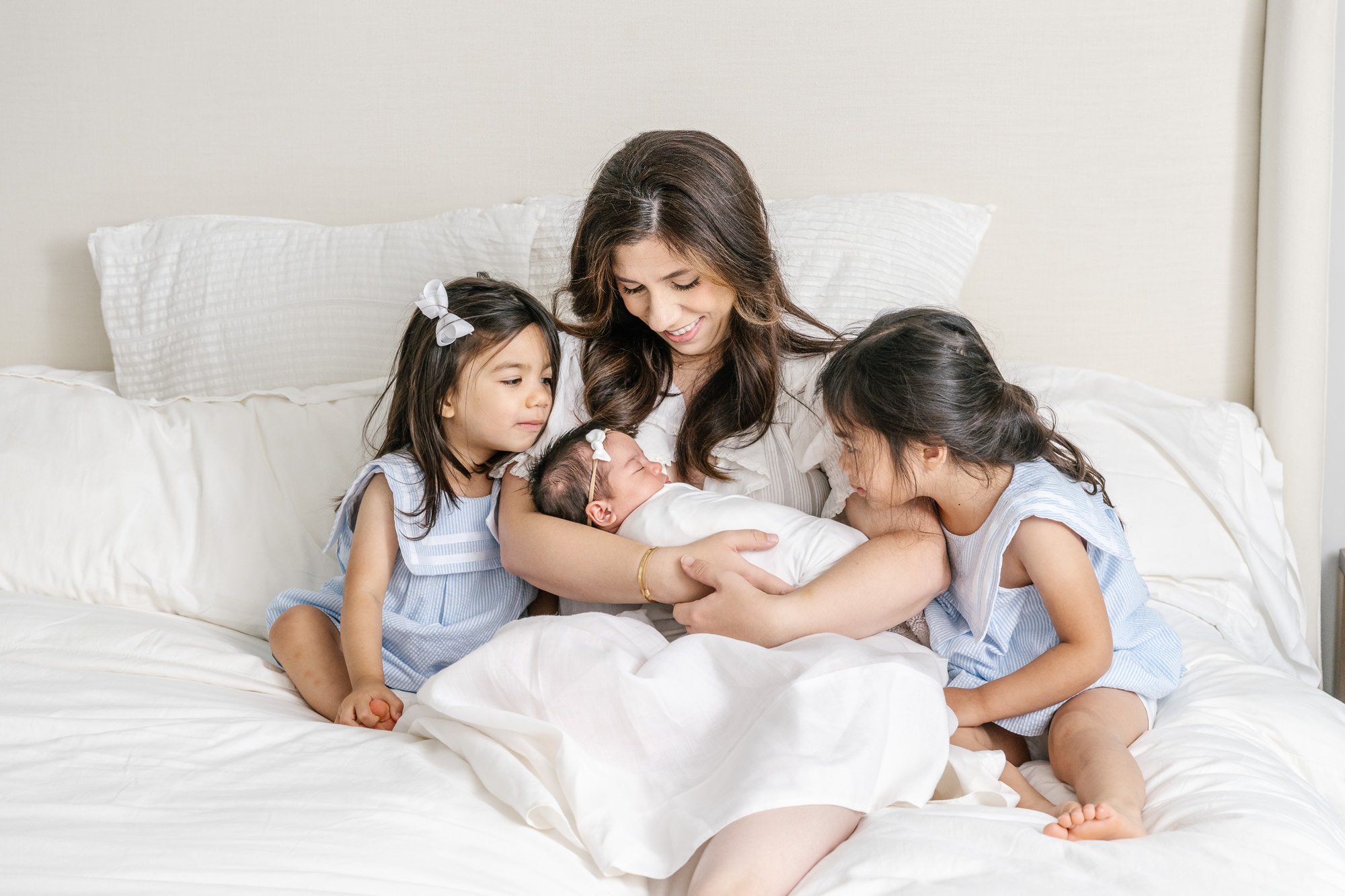  Mom snuggling up in bed holding her newborn daughter while her other two daughters cuddle up next to her and baby sister. #newyorkcityphotographer #familyportraits #newbornportraitsession #inhomephotography #familyoffive #familyposeinspo #newjerseyf