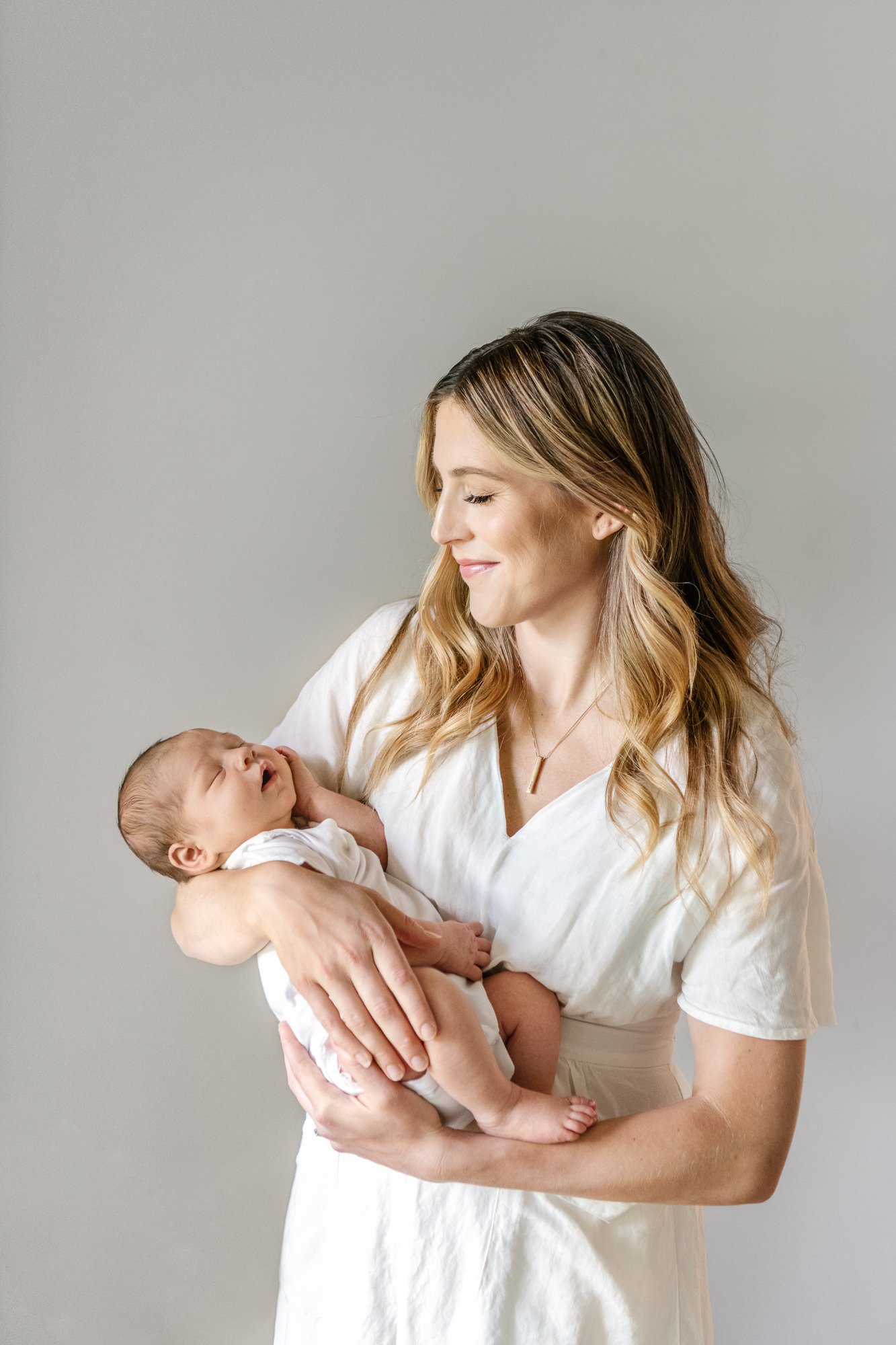  Mom wearing a casual white dress holds her newborn baby boy in her arms while snuggling him close in an affectionate and candid photo. #candidposeinspo #newbornphotography #shorthillsphotographer #familyphotographersinnewjersey #inhomeportraits #new