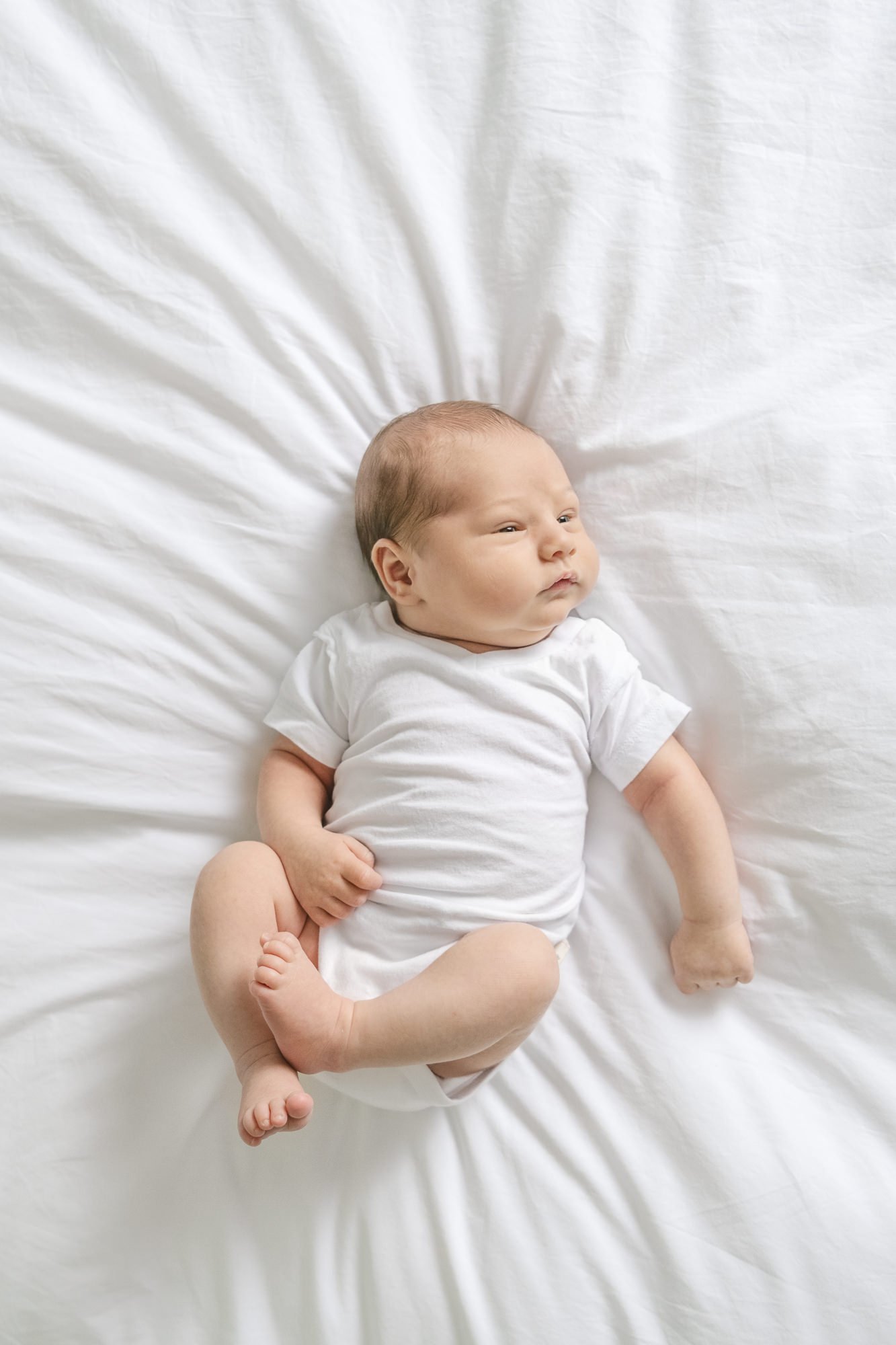  Newborn baby boy lays on a white down comforter while wearing a simple white onesie during his newborn portrait session. #candidposeinspo #newbornphotography #shorthillsphotographer #familyphotographersinnewjersey #inhomeportraits #newbornposeinspir