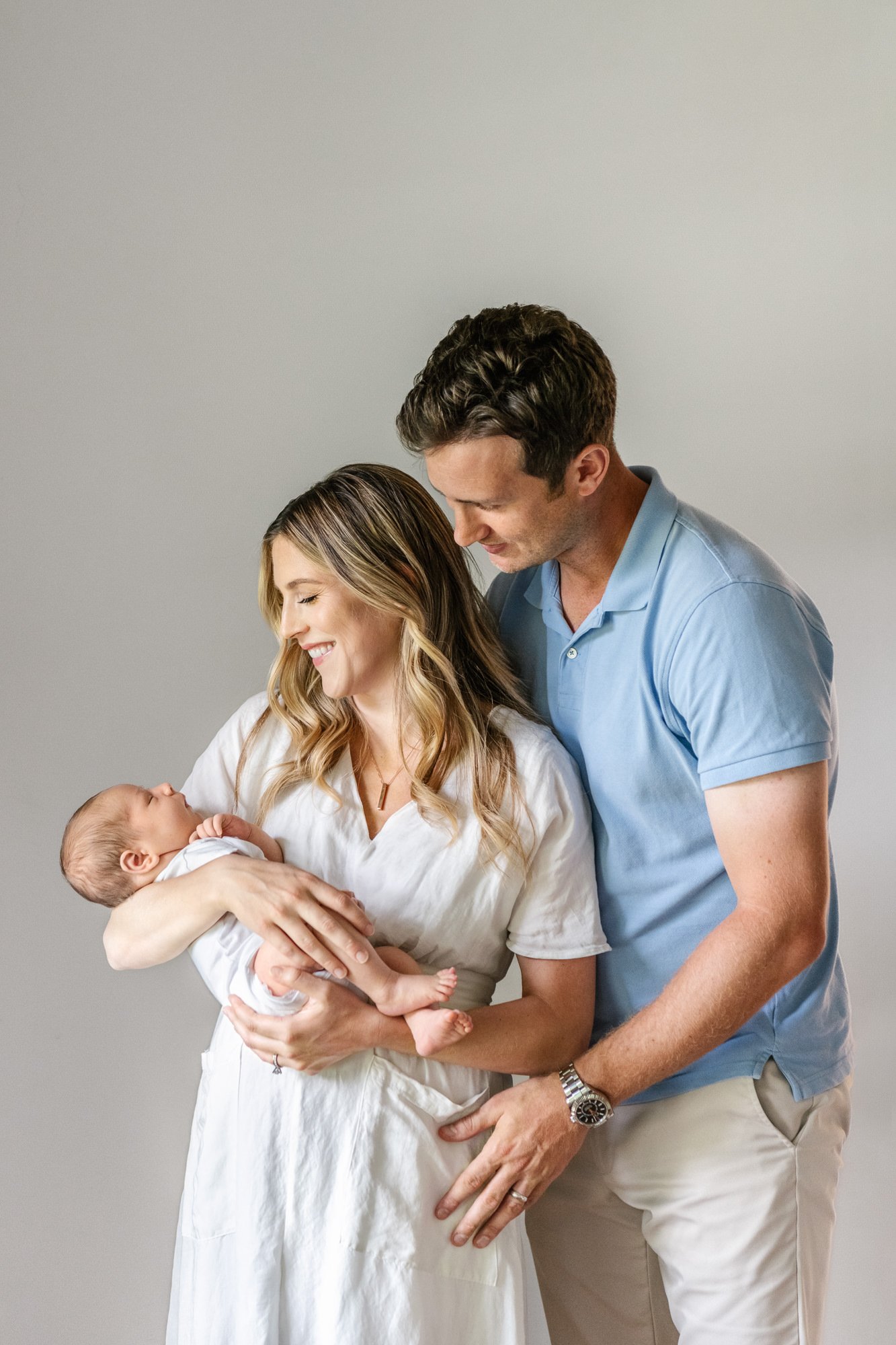  Dad holds mom from behind while both parents smile at their precious newborn baby boy during their in-home portrait session. #candidposeinspo #newbornphotography #shorthillsphotographer #familyphotographersinnewjersey #inhomeportraits #newbornposein