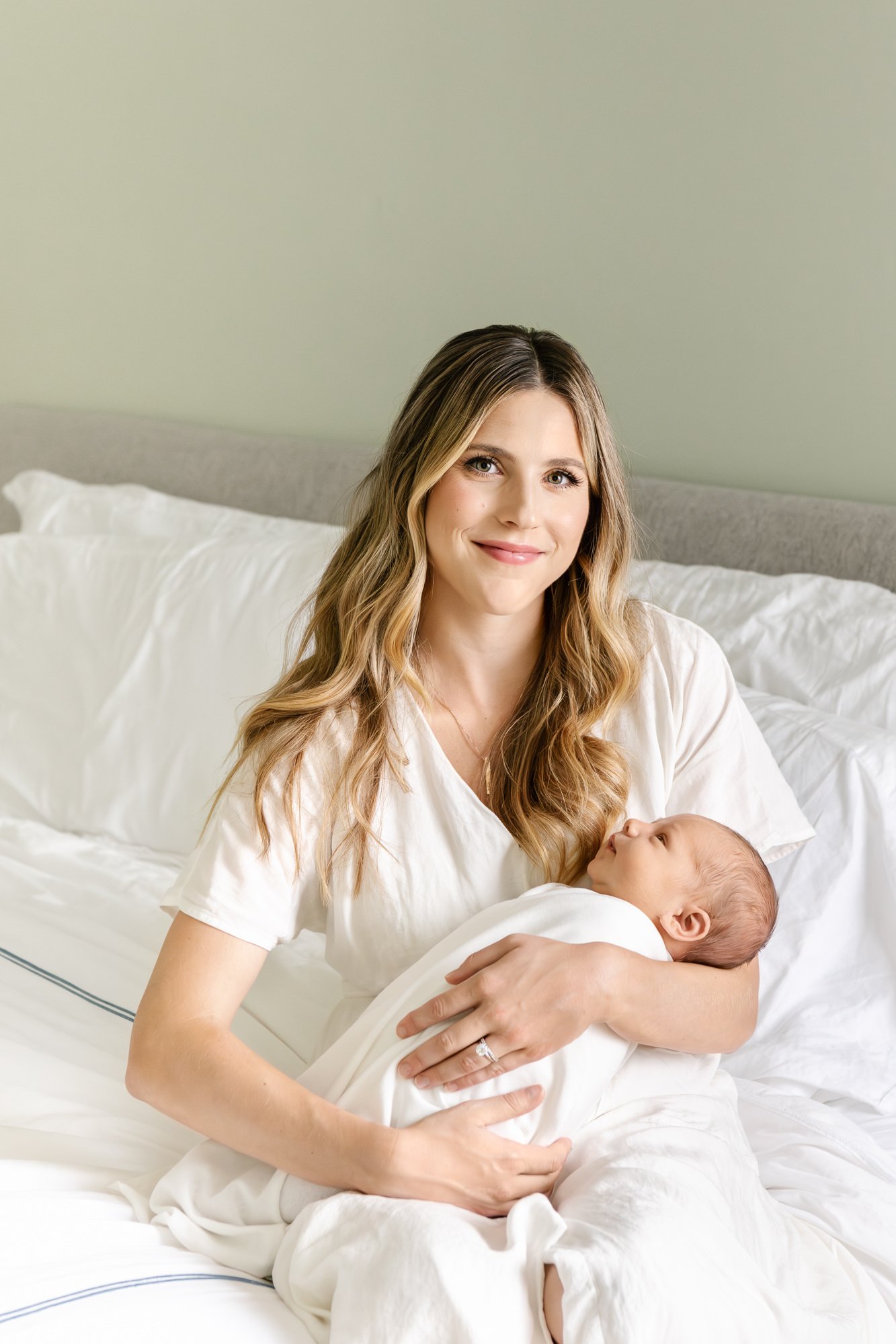  Mom sits on a queen size bed in a casual white dress while holding her precious newborn son in her arms for a natural, candid photo. #candidposeinspo #newbornphotography #shorthillsphotographer #familyphotographersinnewjersey #inhomeportraits #newbo