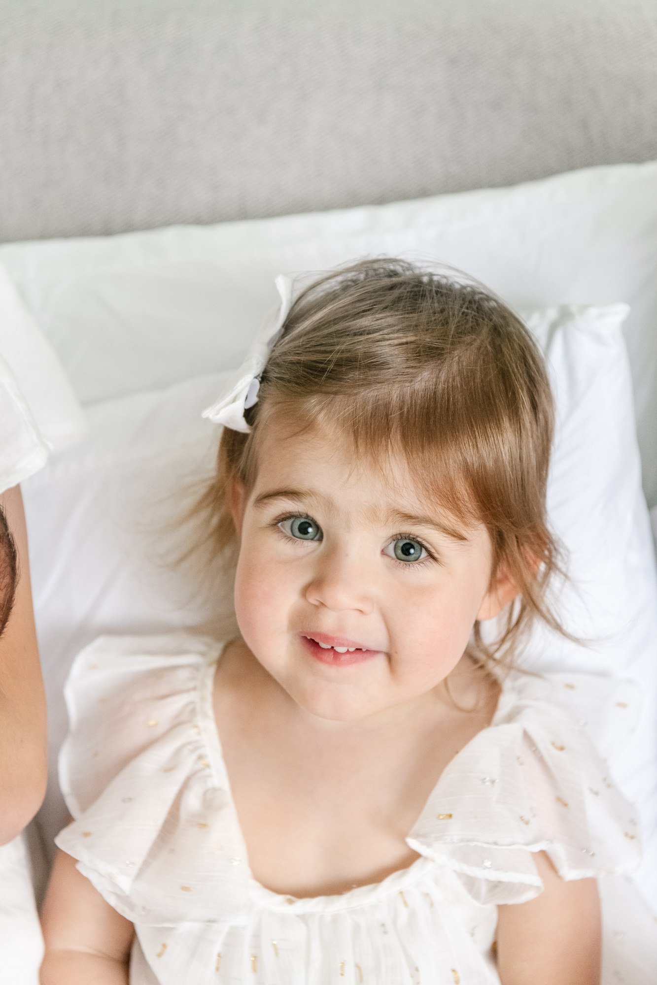  Sweet toddler girl around two years old smiles up at the camera with her beautiful big blue eyes while wearing a feminine white dress. #candidposeinspo #newbornphotography #shorthillsphotographer #familyphotographersinnewjersey #inhomeportraits #new