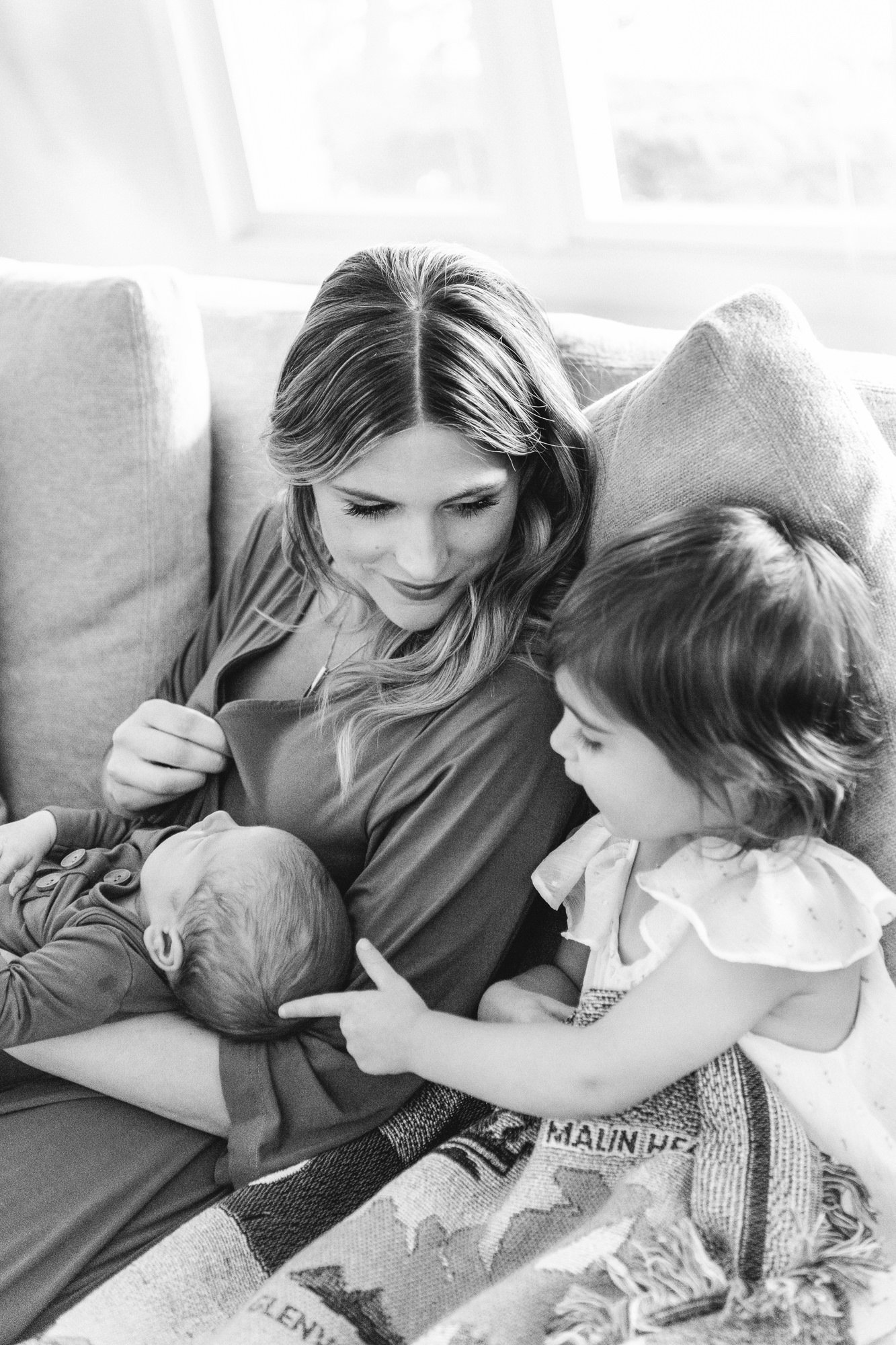  Black and white photo of a mother holding her newborn son while talking with her two year old daughter on the couch. #candidposeinspo #newbornphotography #shorthillsphotographer #familyphotographersinnewjersey #inhomeportraits #newbornposeinspiratio