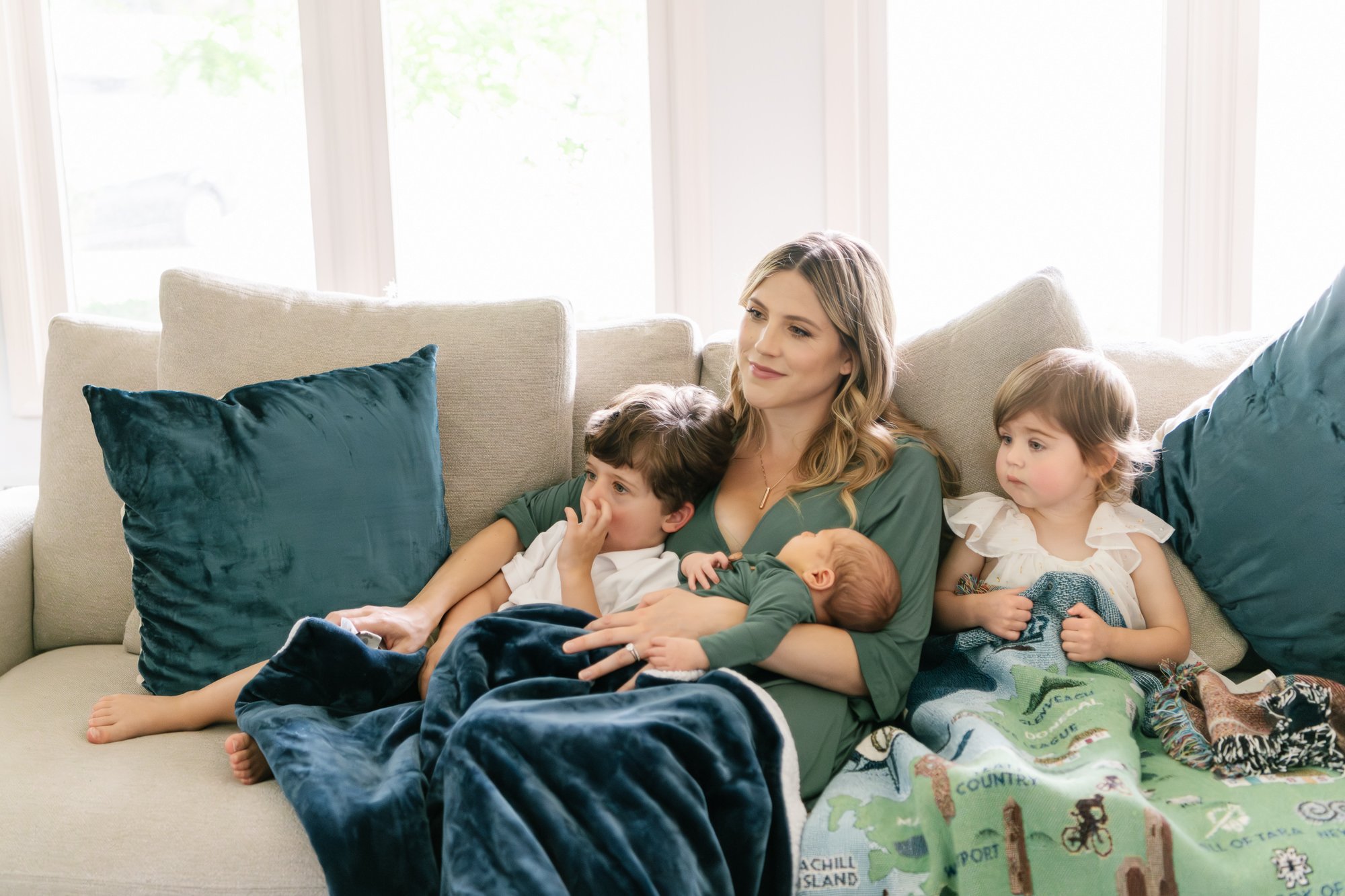  Mother snuggles on the couch with her toddler son and daughter as well as their newborn baby brother to talk and watch a movie as a family. #candidposeinspo #newbornphotography #shorthillsphotographer #familyphotographersinnewjersey #inhomeportraits