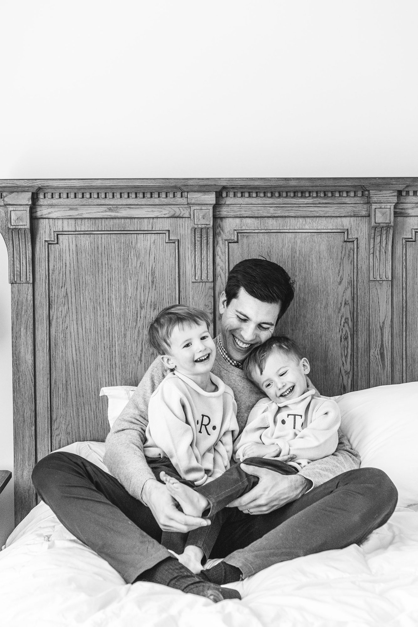   Black and white in-home family photos with dad sitting on bed holding two sons, smiling at the camera. Family photoshoot pose ideas in-home family portrait pose ideas #EssexCountyFamilyPhotographer #MorrisCountyFamilyPhotographer #ManhattanFamilyPh
