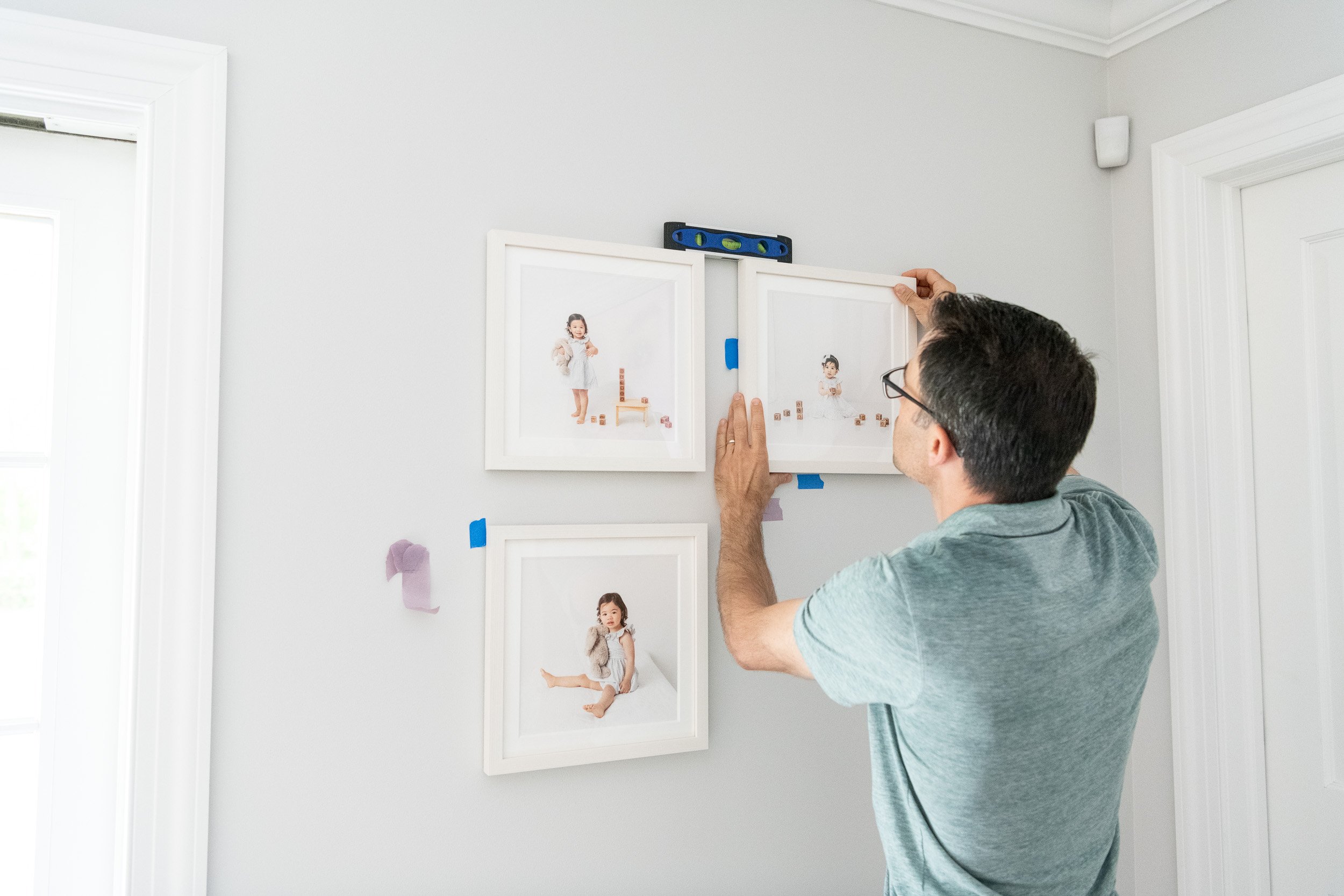   Dad carefully hanging gallery wall of framed photos of toddler playing with wood toys in nursery. Nursery gallery wall inspiration #EssexCountyFamilyPhotographer #PassaicCountyFamilyPhotographer #NorthernNewJerseyFamilyPhotographer #ManhattanPhotog