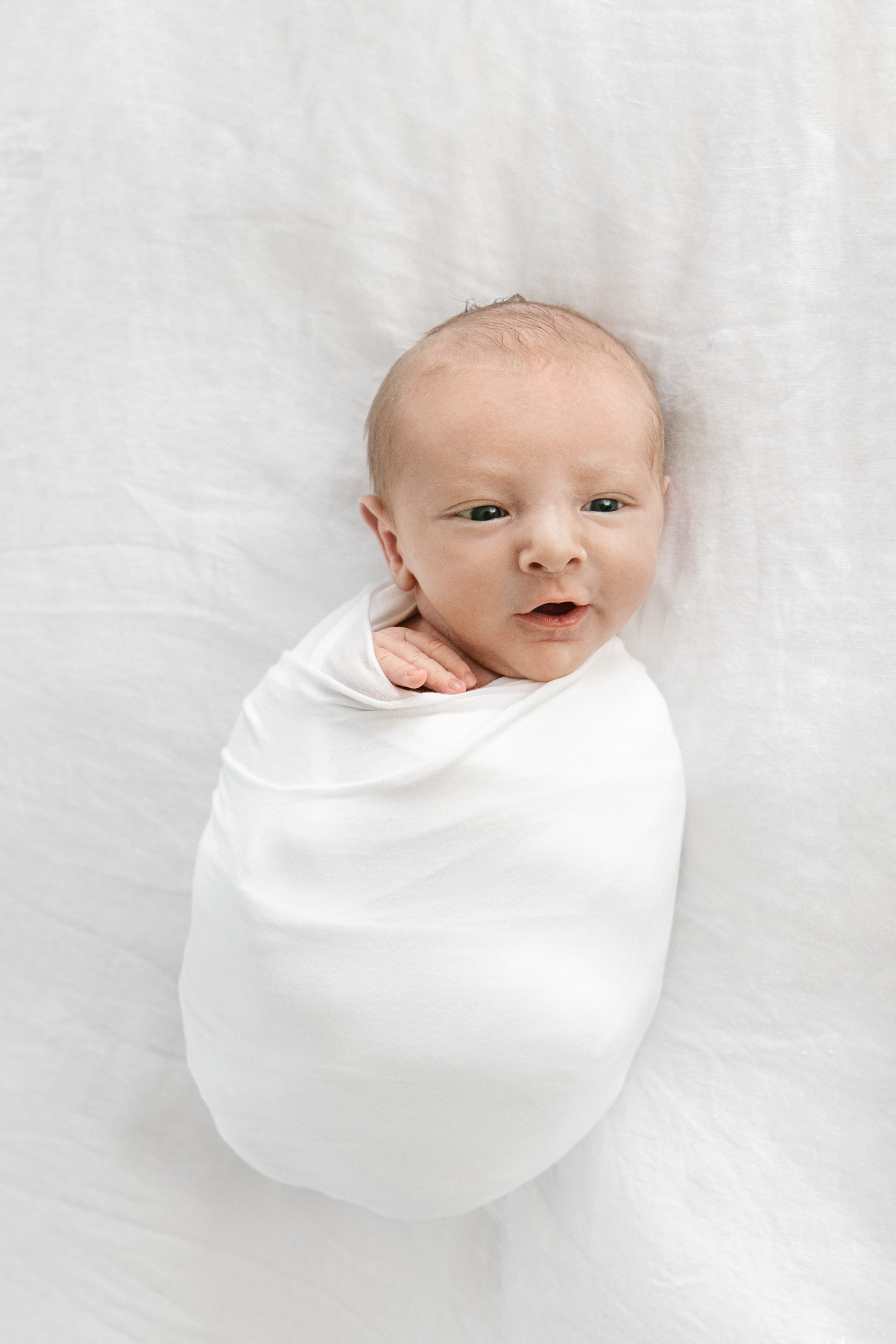   Newborn baby wrapped up in white swaddle blanket, smiling at the camera in at-home portrait taken by Nicole Hawkins, a newborn photographer in northern New Jersey. #EssexCountyFamilyPhotographer #PassaicCountyFamilyPhotographer #NorthernNewJerseyFa