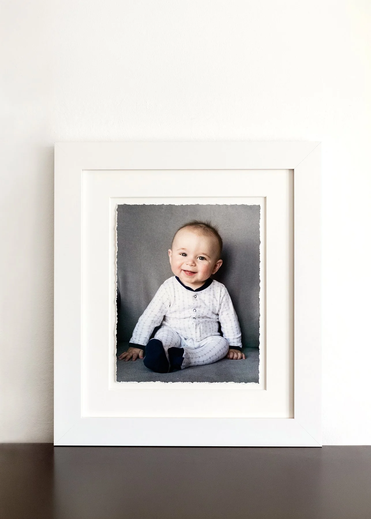 A framed photograph of a smiling 6 month old baby wearing a white outfit and sitting on a gray surface.