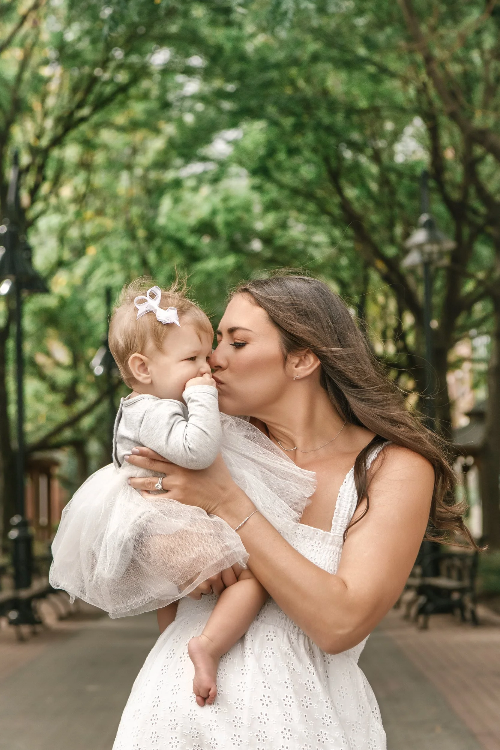 A woman holding and kissing her 6 month baby in a Hoboken park, with trees lining the walkway in the background. 