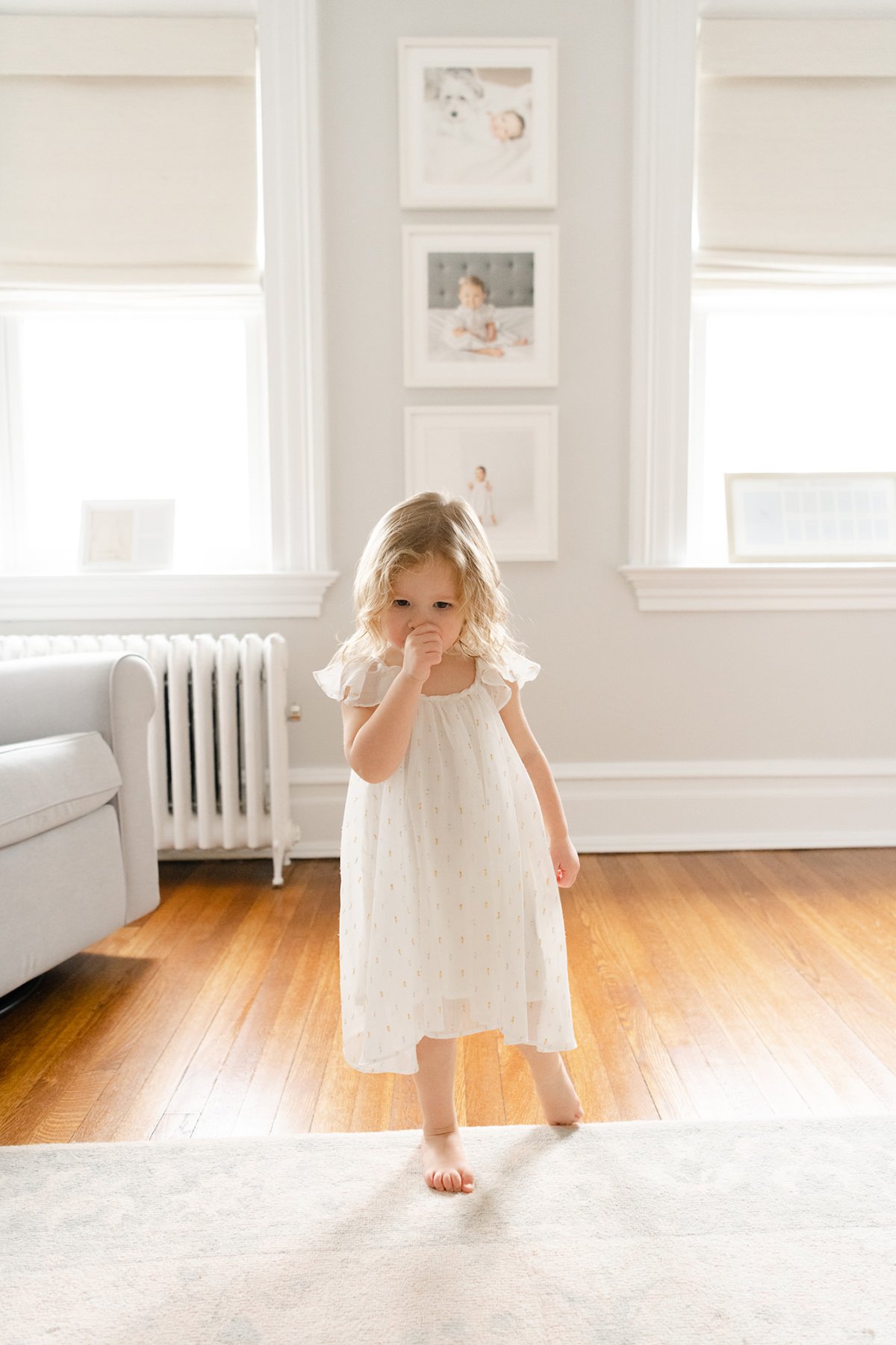 Toddler with blonde hair in a white dress standing barefoot on a wooden floor in a brightly lit Chatham home in front of framed newborn, 6 month, and first birthday portraits.