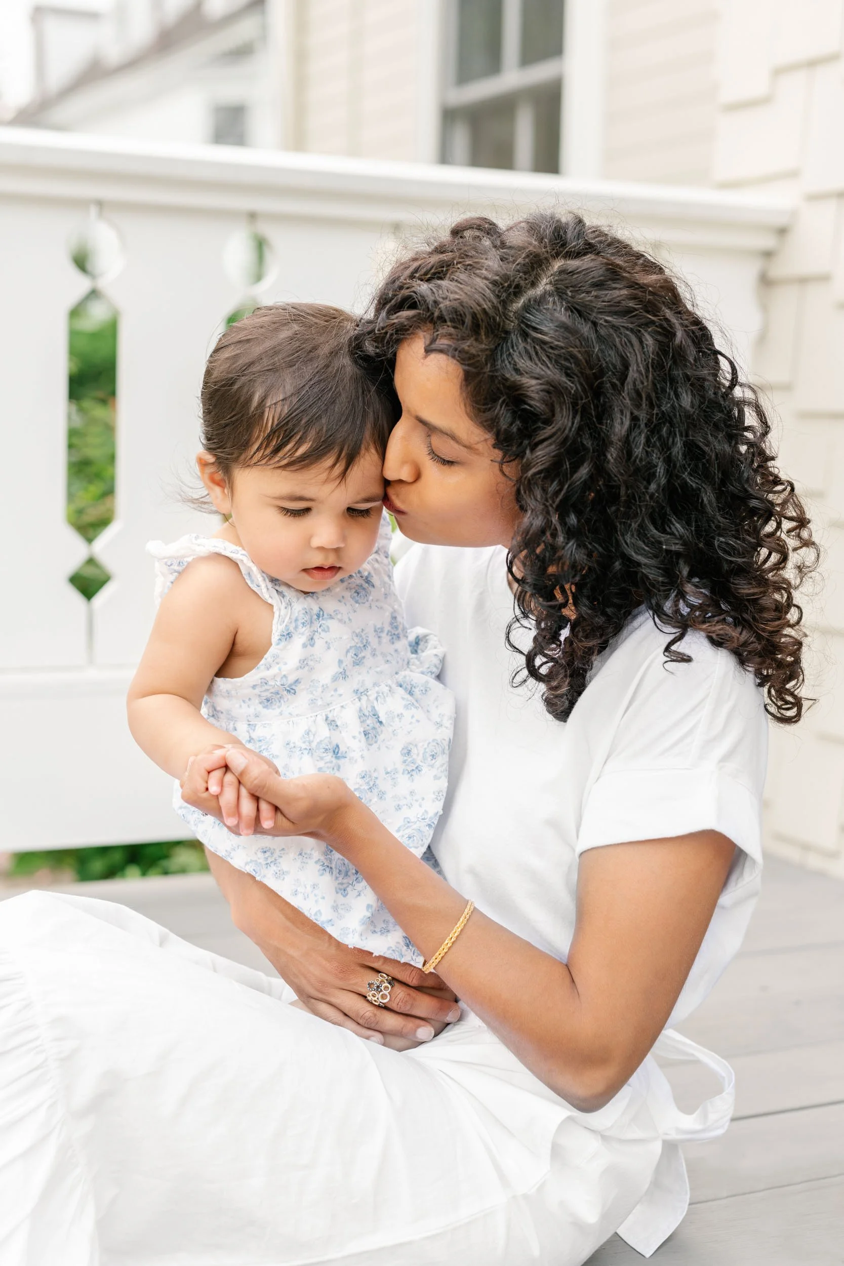 A woman with curly hair kisses a young girl on the forehead while holding her close, sitting at home in Glen Ridge NJ backyard.