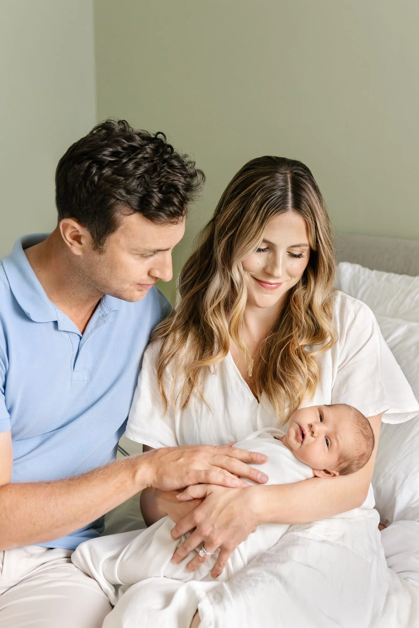 A mother and father sitting on their bed at home in Short Hills NJ holding their newborn baby.