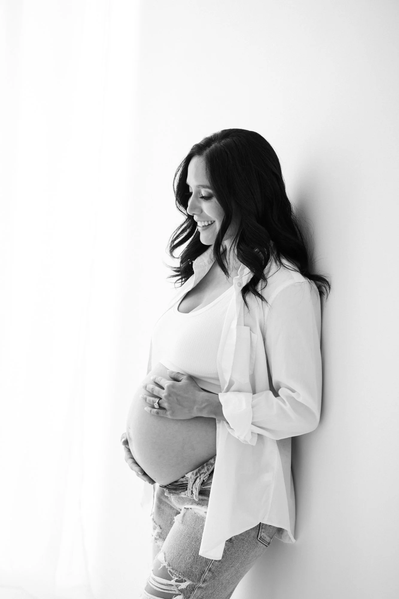 A black-and-white photograph of a pregnant woman standing and smiling, leaning against a plain white wall, cradling her belly with both hands, wearing distressed jeans and a loose shirt layered over a tank top.