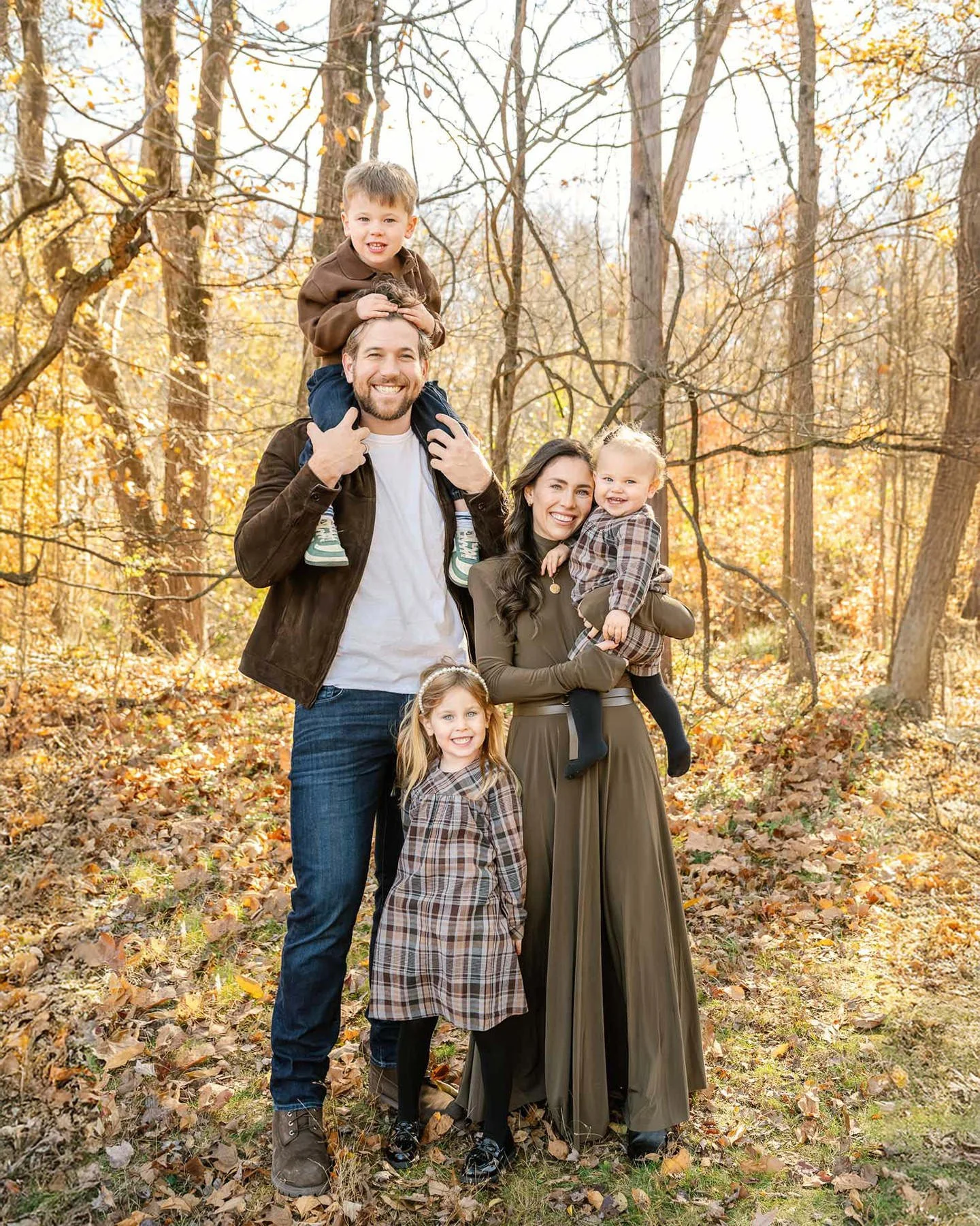 Family of five smiling in a fall forest, with trees and fallen leaves.