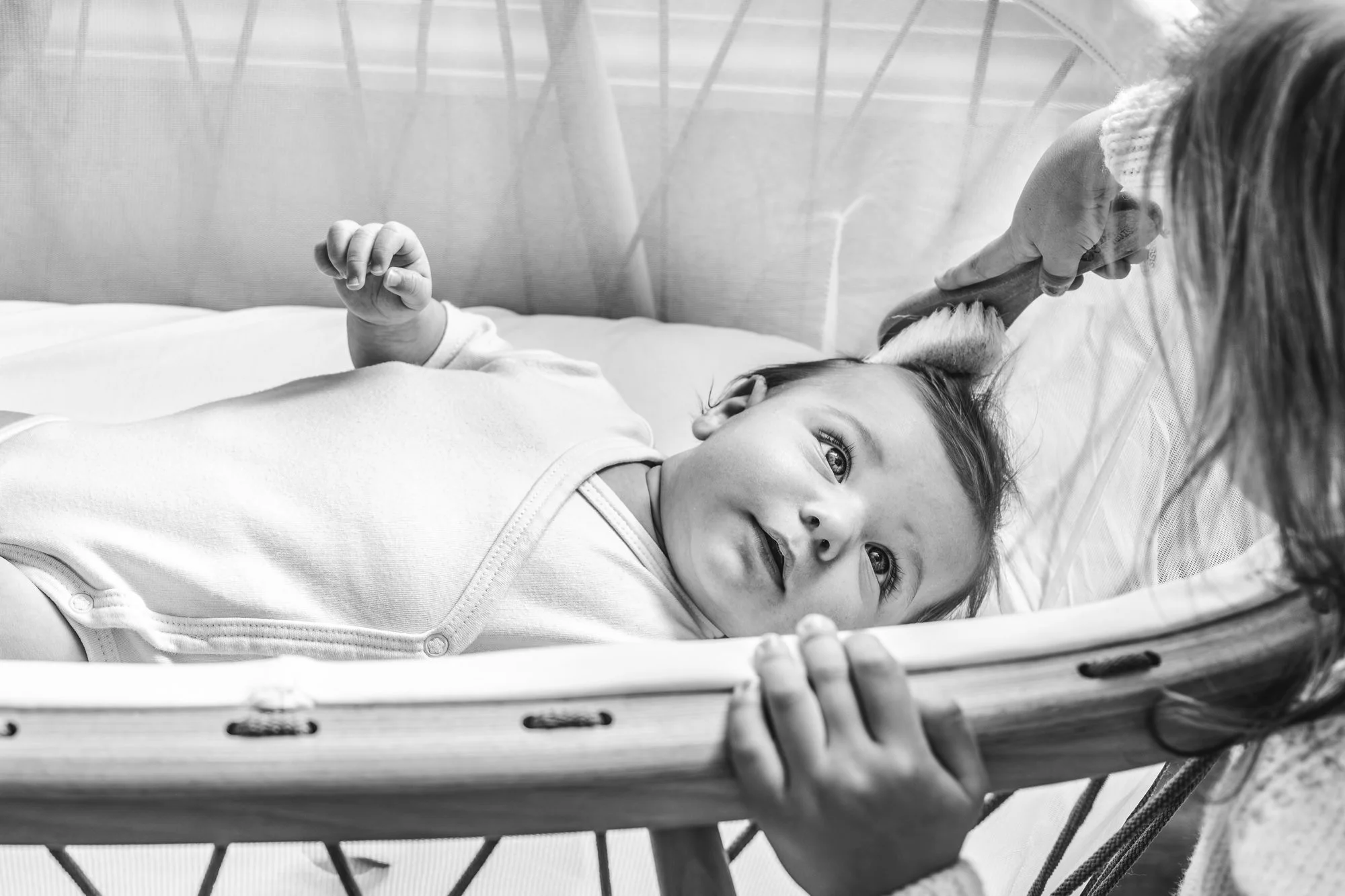Three month old baby lying in a crib, smiling and looking up, as big sister gently brushes hair from the baby's forehead.