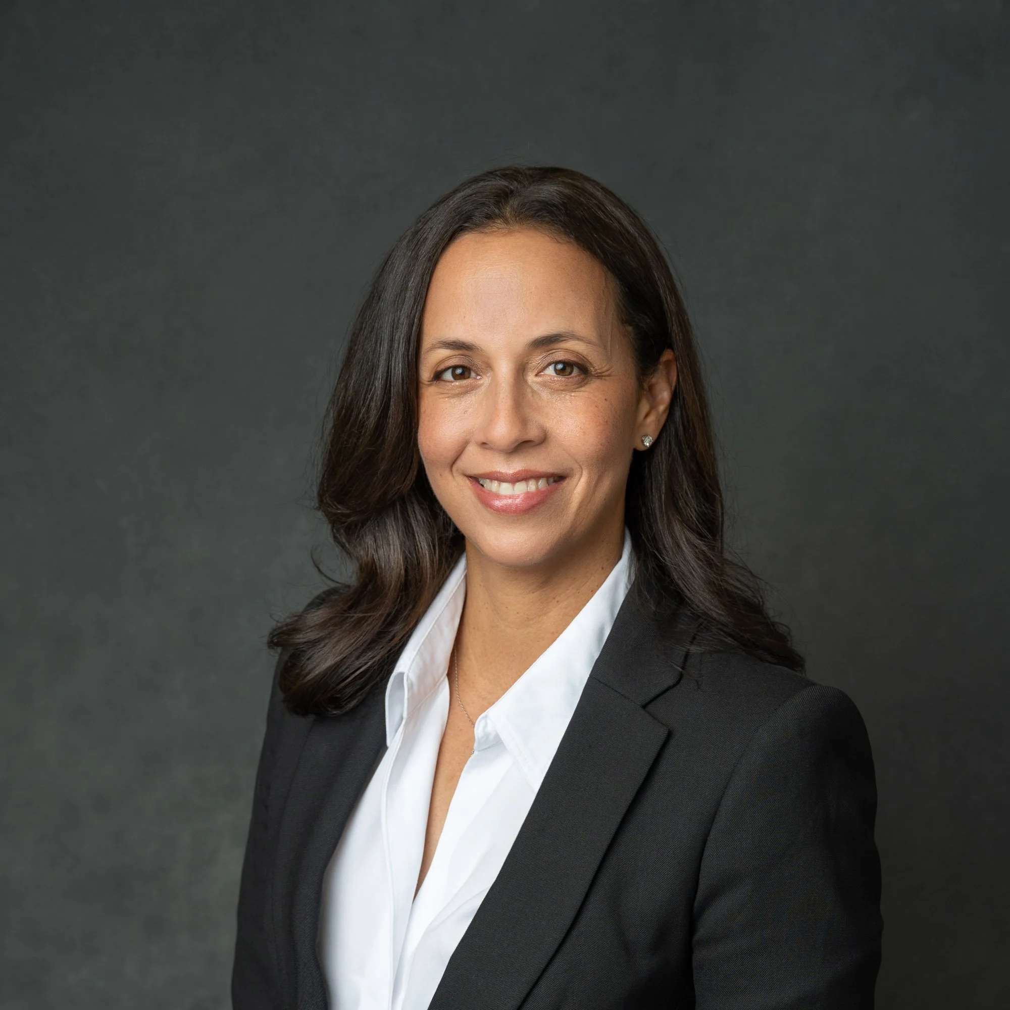 Professional headshot portrait of a latina finance VP with shoulder length dark hair, smiling, dressed in a black blazer and white shirt, against a dark gray background.