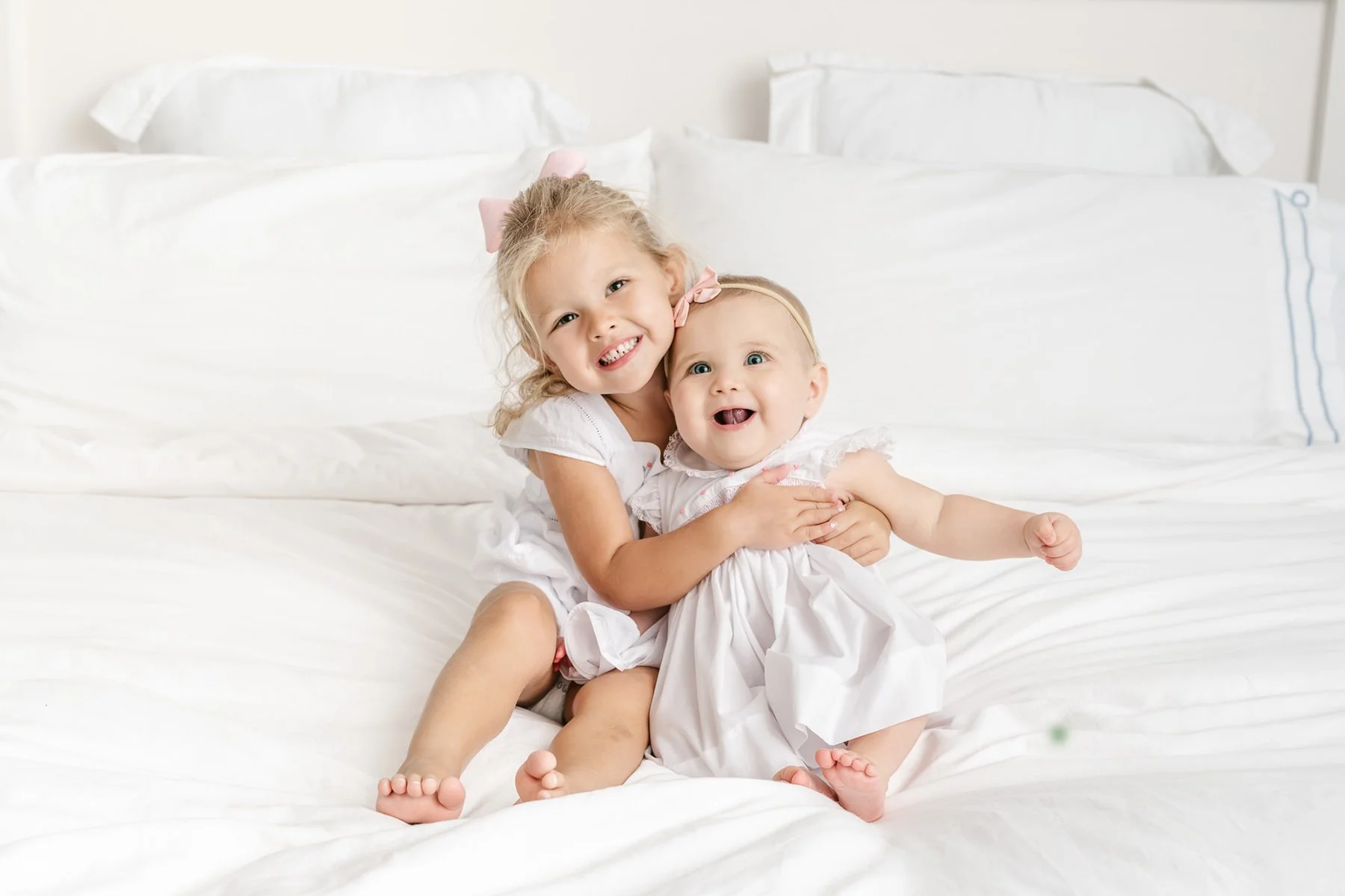 Two young sisters in white dresses sitting on a white bed, smiling at the camera.