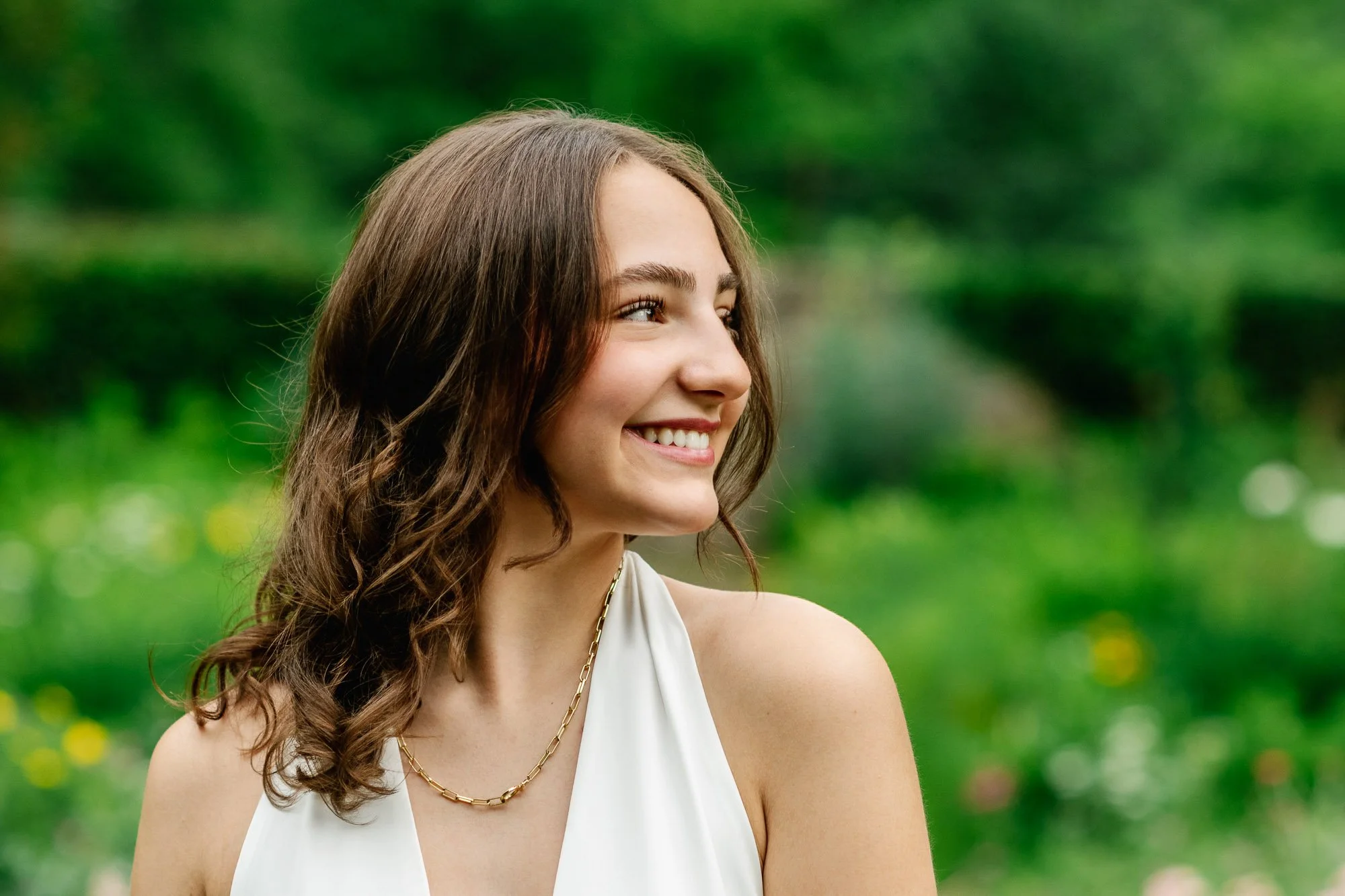 Profile of a female high school senior with long, curly brown hair smiling outdoors, wearing a white sleeveless top and a gold chain necklace, with a blurred green natural background.