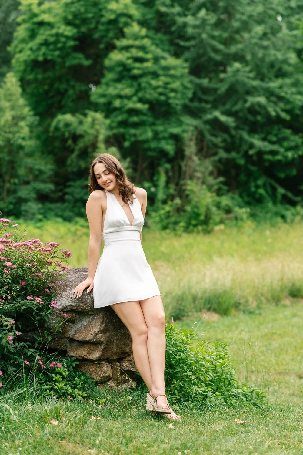 Painterly outdoor senior portrait of girl in white satin dress leaning against a stone wall and wildflowers at Cross Estate Gardens