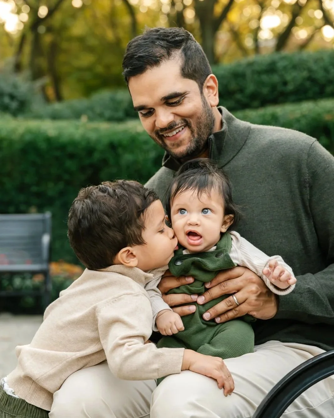 Father with dark hair sitting on NJ park bench holding his baby on his lap while dark haired toddler son wearing cream clothing kisses baby brother.