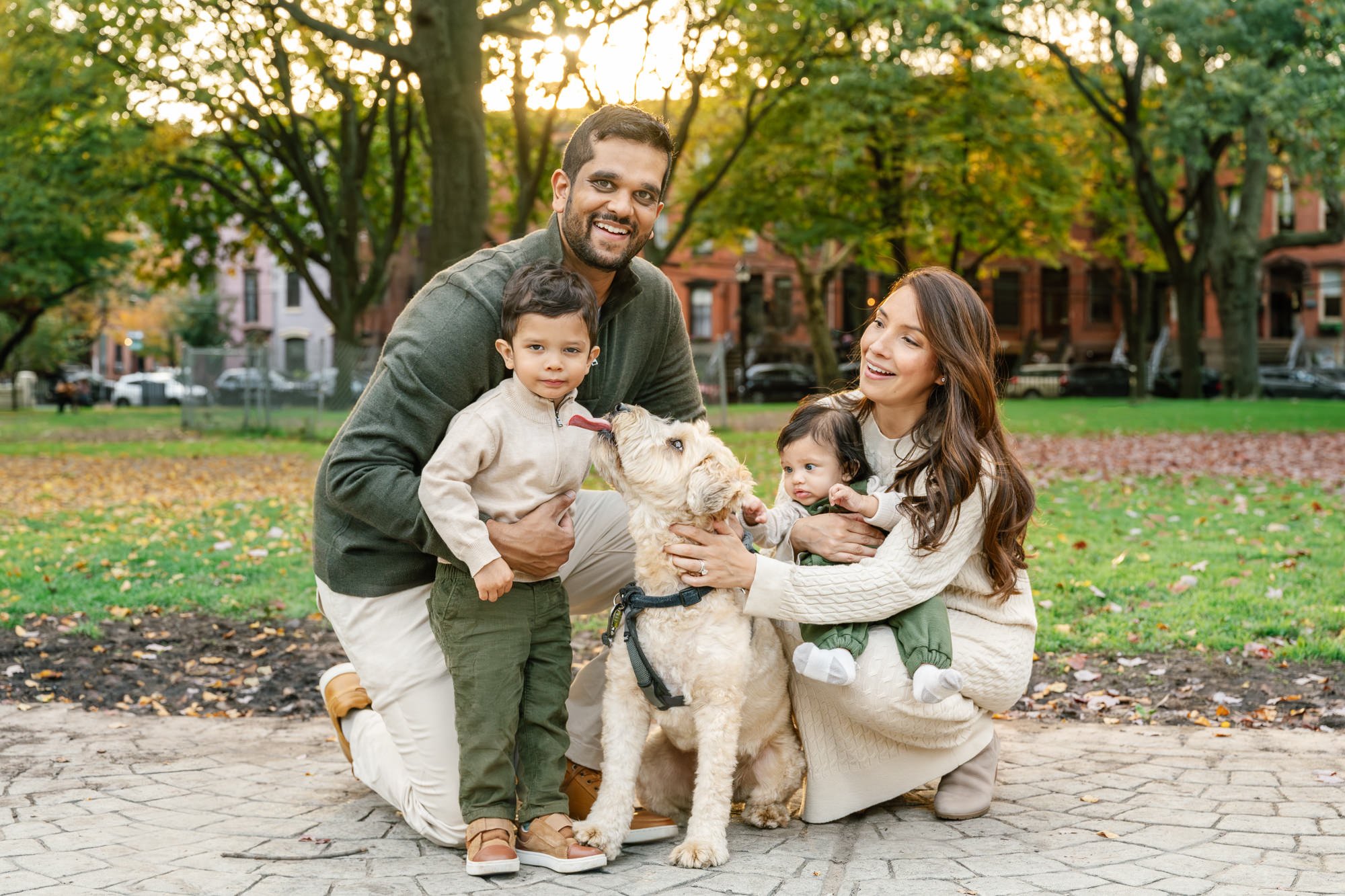A family of four, including two young children and a dog, enjoying time together in a park during autumn with colorful fall leaves and trees in the background.