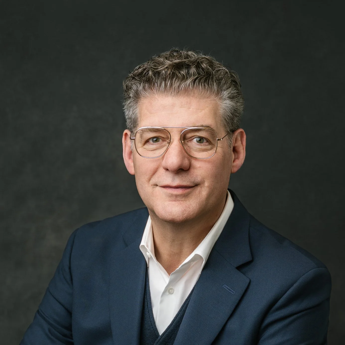 Classic headshot of a middle-aged man with short, curly gray hair, wearing glasses, a white shirt, and a dark blue blazer, smiling against a dark background.
