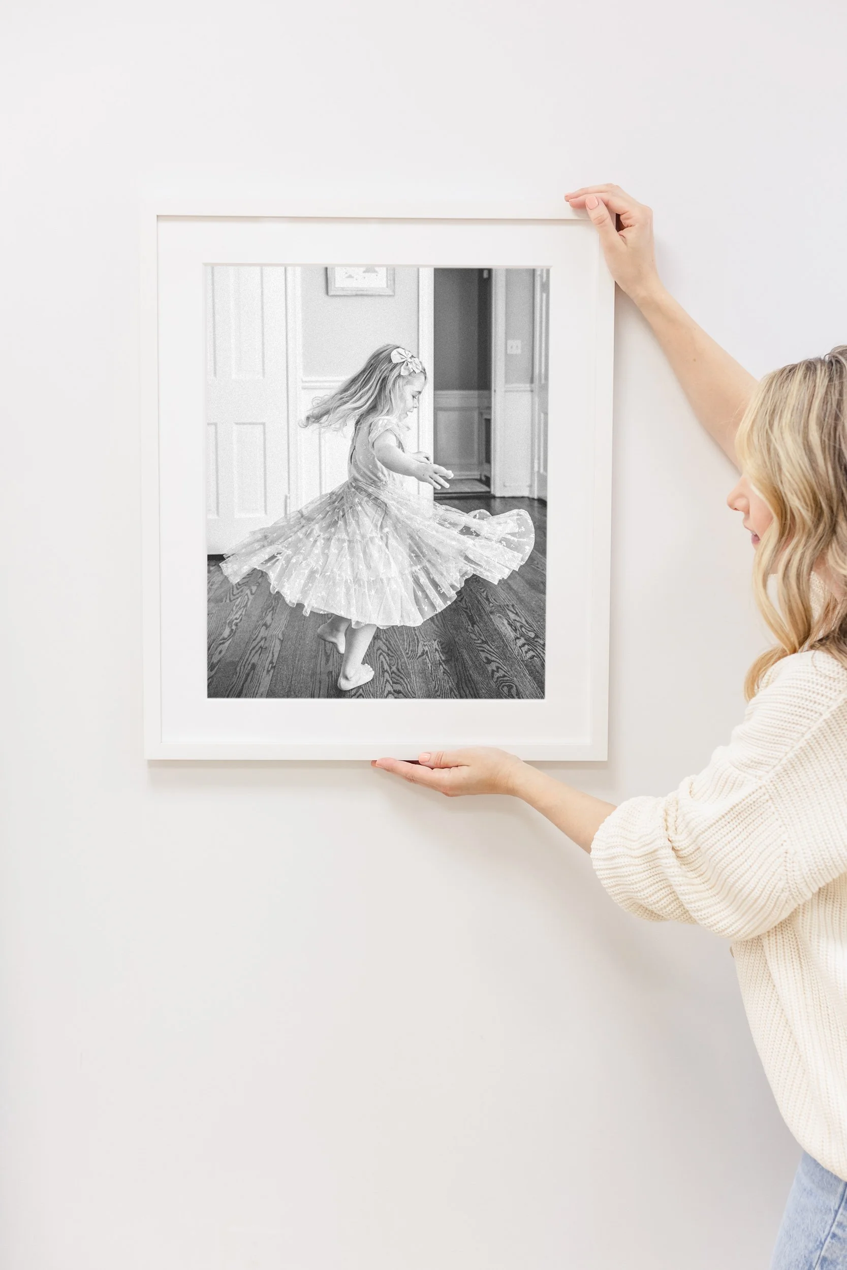 A woman hanging a black and white photograph of a young girl twirling in a dress on a white wall.
