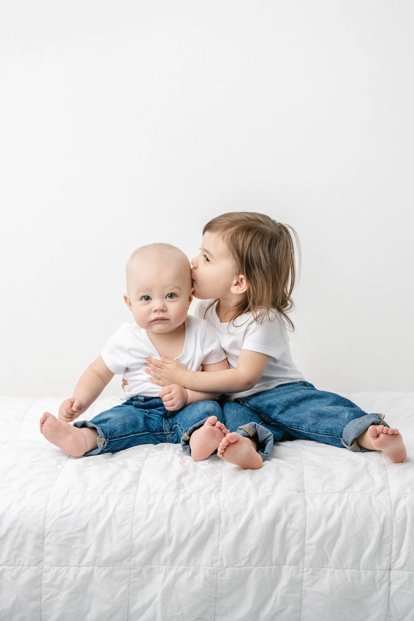 Young sisters wearing jeans sitting on a white bed against a plain white wall, with older sister kissing one year old baby's head.