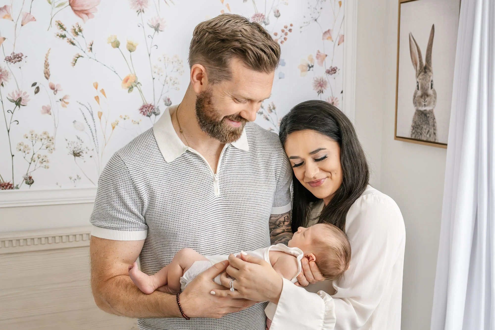 New mom and dad smile down at their newborn daughter during an in-home portrait session by Nicole Hawkins photography. Couple stands in a classic nursery. Neutral outfit inspiration. #centralNewJerseyphotographer #inhomeportraits #newbornphotoshoot #