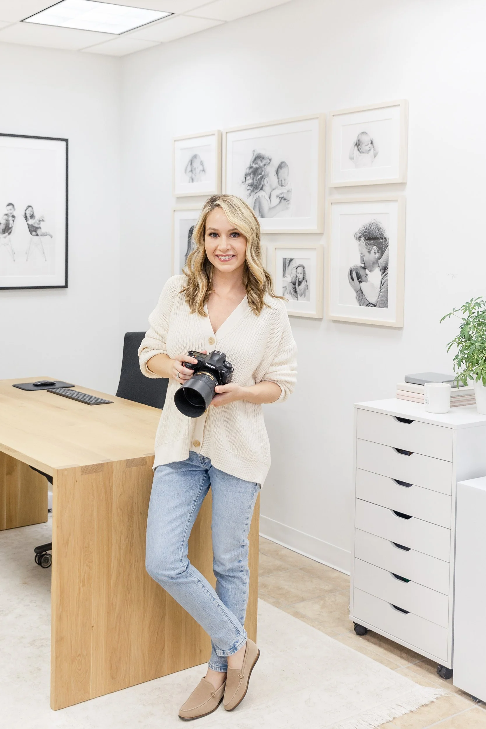 Professional photographer with blonde hair holding Nikon camera, wearing a cream-colored cardigan and light blue jeans, stands in a modern, minimalist family photo studio in Maplewood nj.