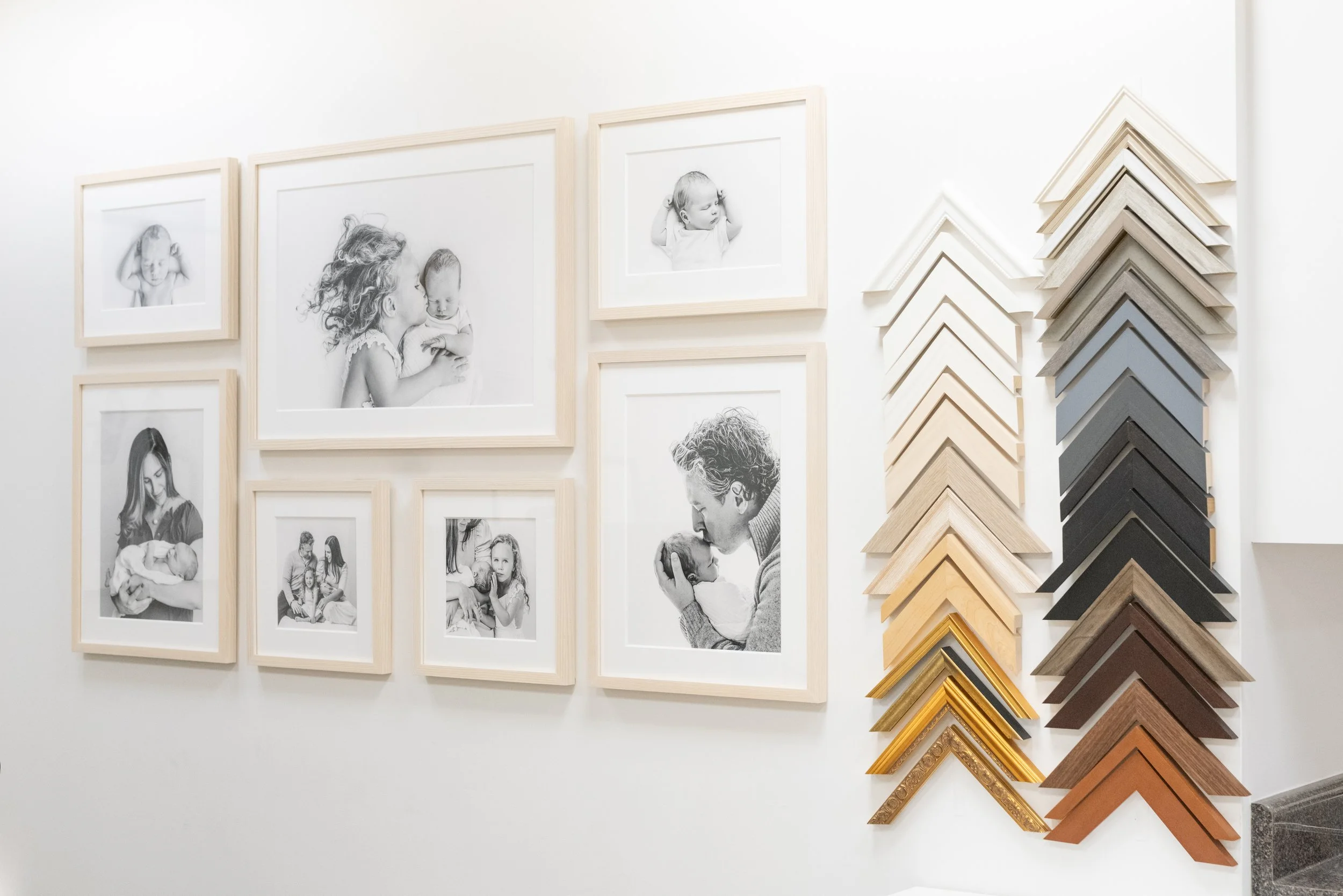 Gallery wall showcasing custom framed black-and-white newborn photographs and a display of picture frame corners.