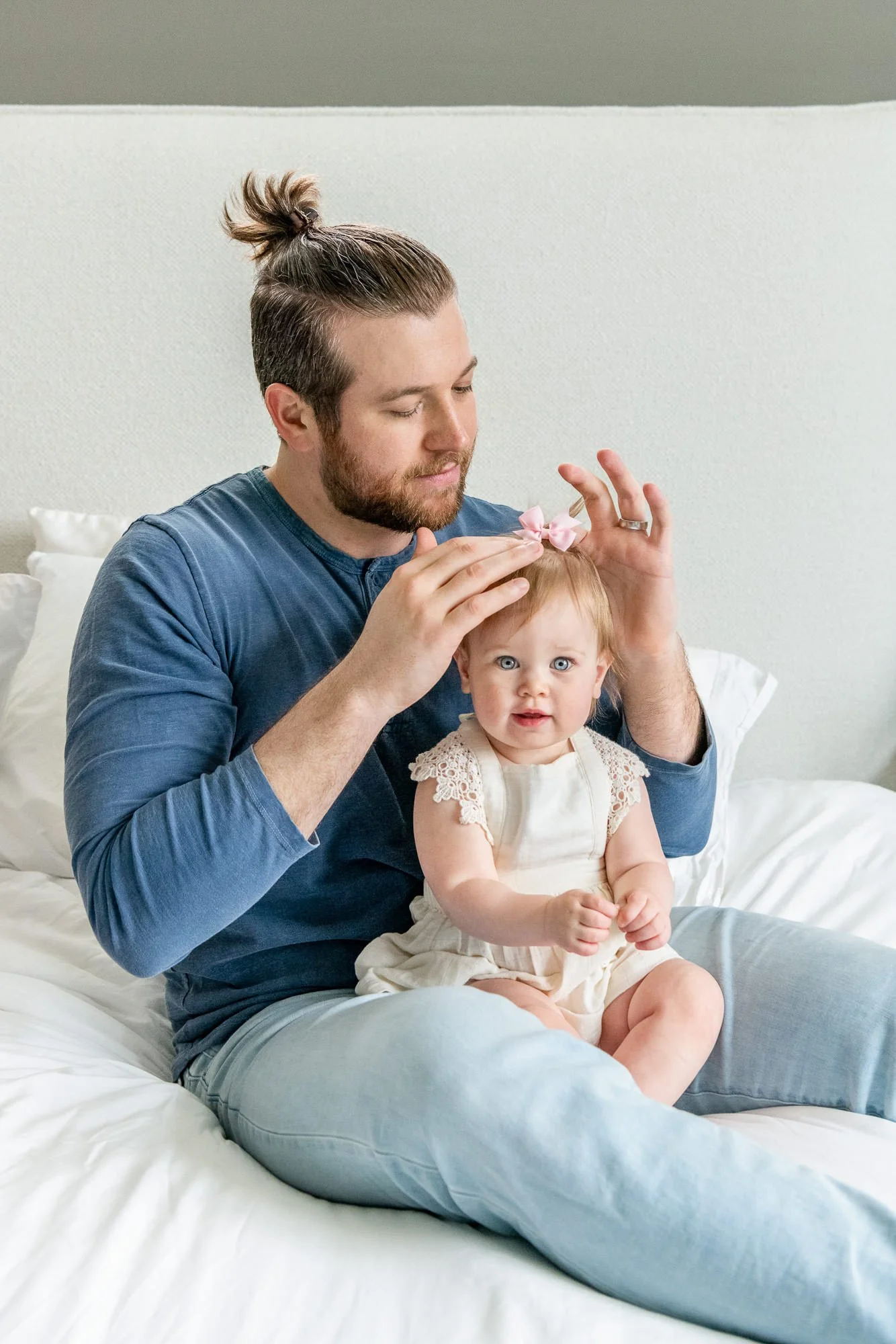 A  father with a beard and bun, wearing a blue shirt, sitting on his bed brushing his young daughters hair before newborn photography session in their Closter home.