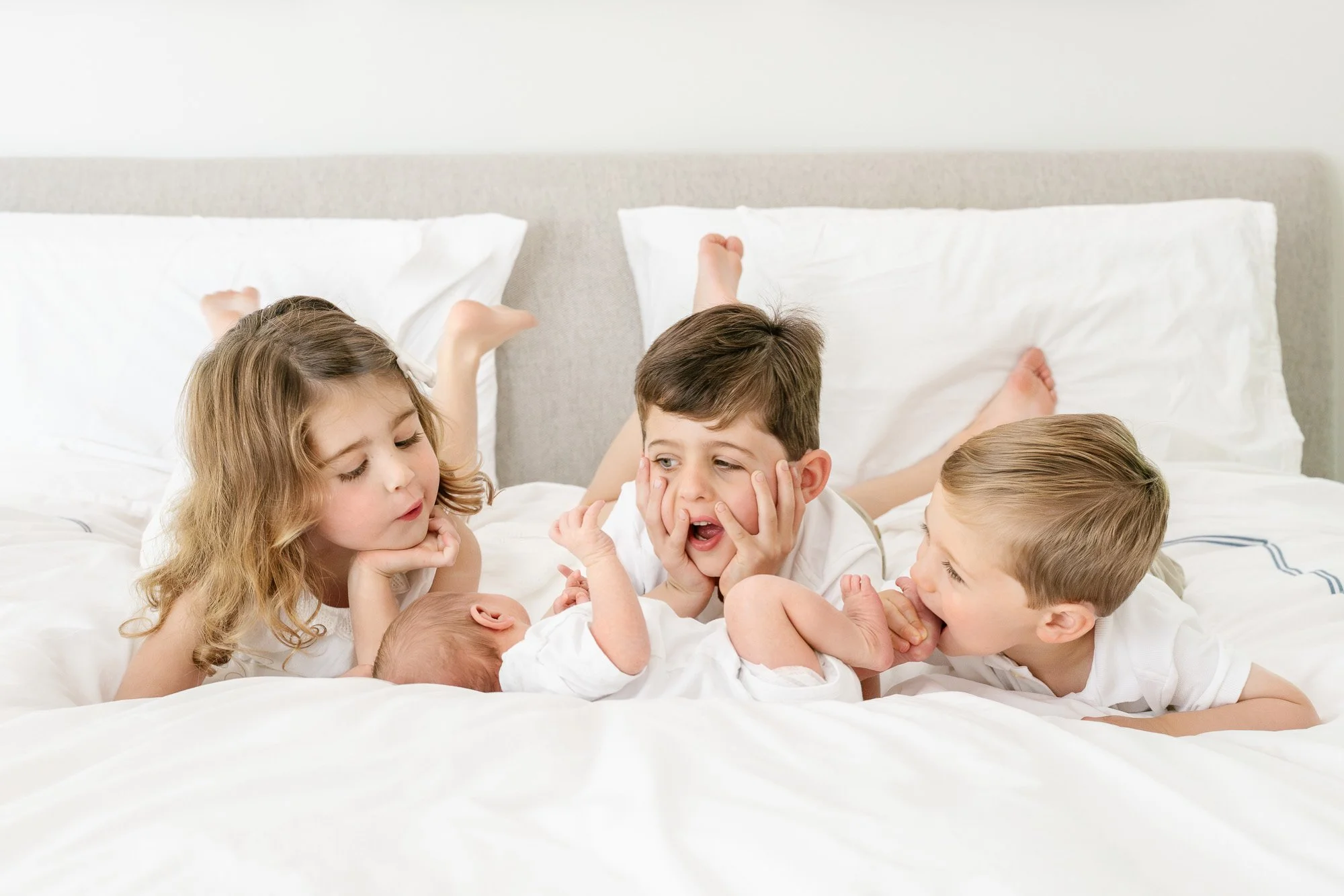 Three young siblings playfully interacting with their newborn baby brother on a white bed in their Short Hills, NJ home.