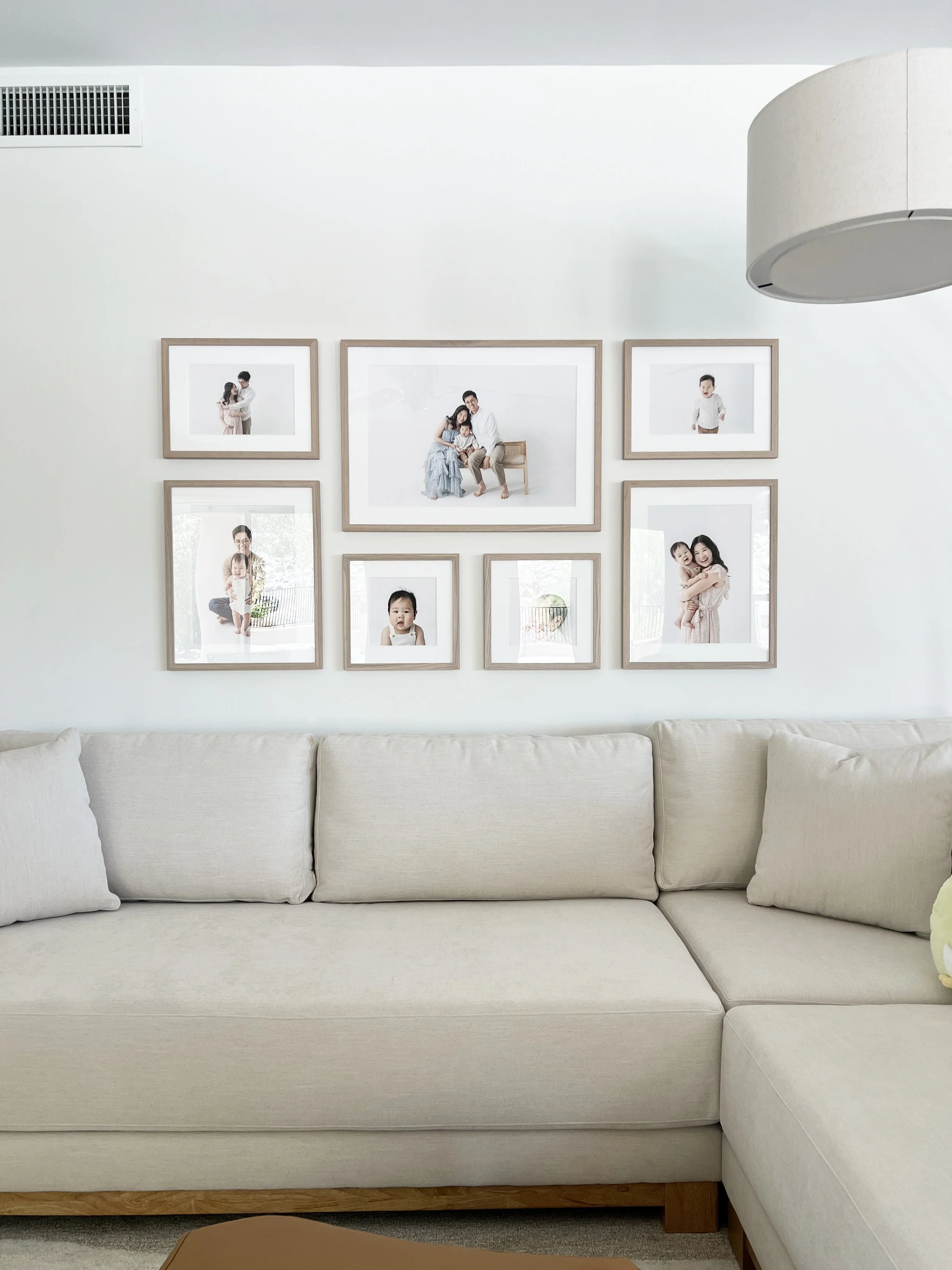 A white living room wall decorated with a gallery of seven framed family photos, above a beige sofa with matching cushions.