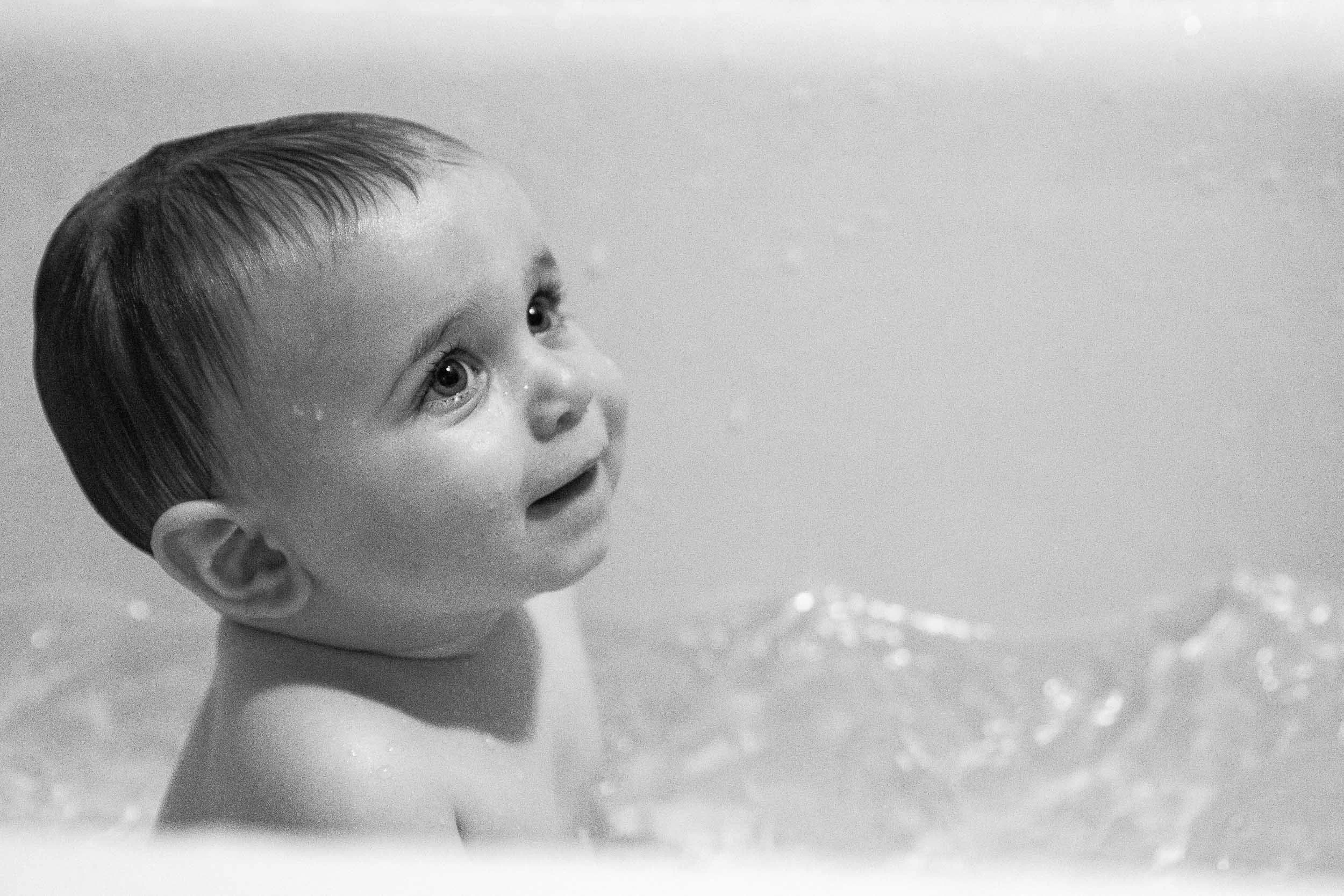Black and white photo of baby at home in a bathtub with wet hair looking upwards with a joyful expression.