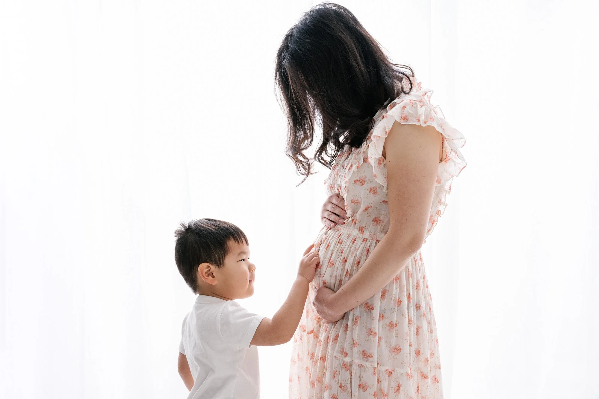 Bright studio maternity portrait of three year old boy gently touching moms pregnant belly. Mom with black hair wears a cream and soft pink flowered dress.