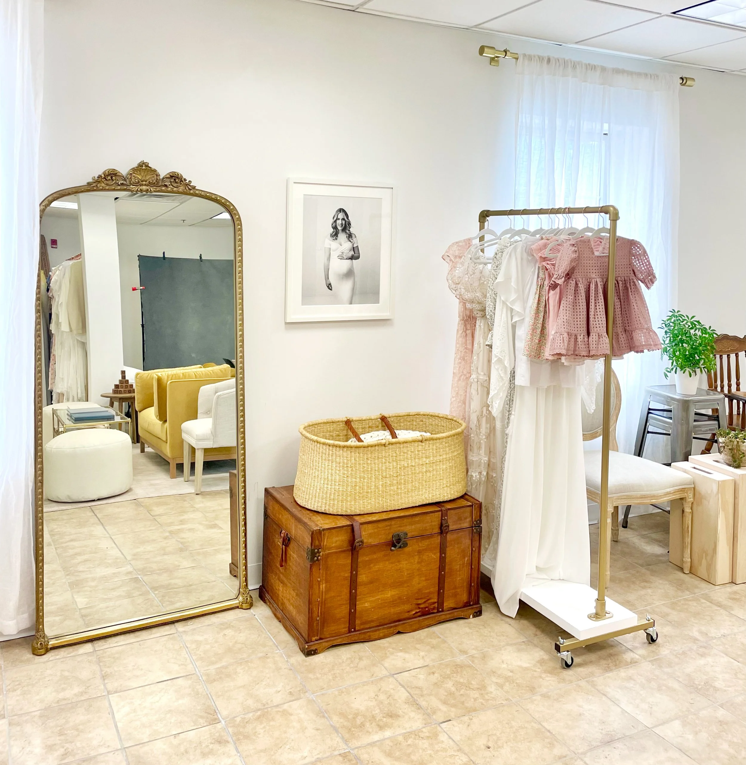 A fitting room area in family portrait studio area with a large mirror, a clothing rack with pink and white dresses, a wooden chest, and a white curtain.