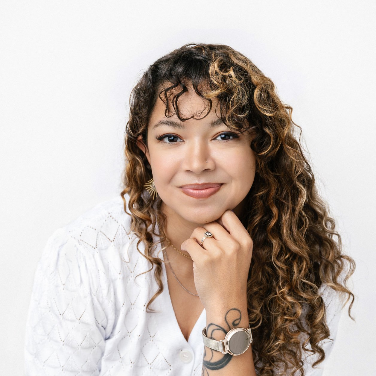 A woman with curly hair and light brown highlights, wearing a white knit blouse rests her chin on her hand, displaying a watch and rings, against a white studio background.
