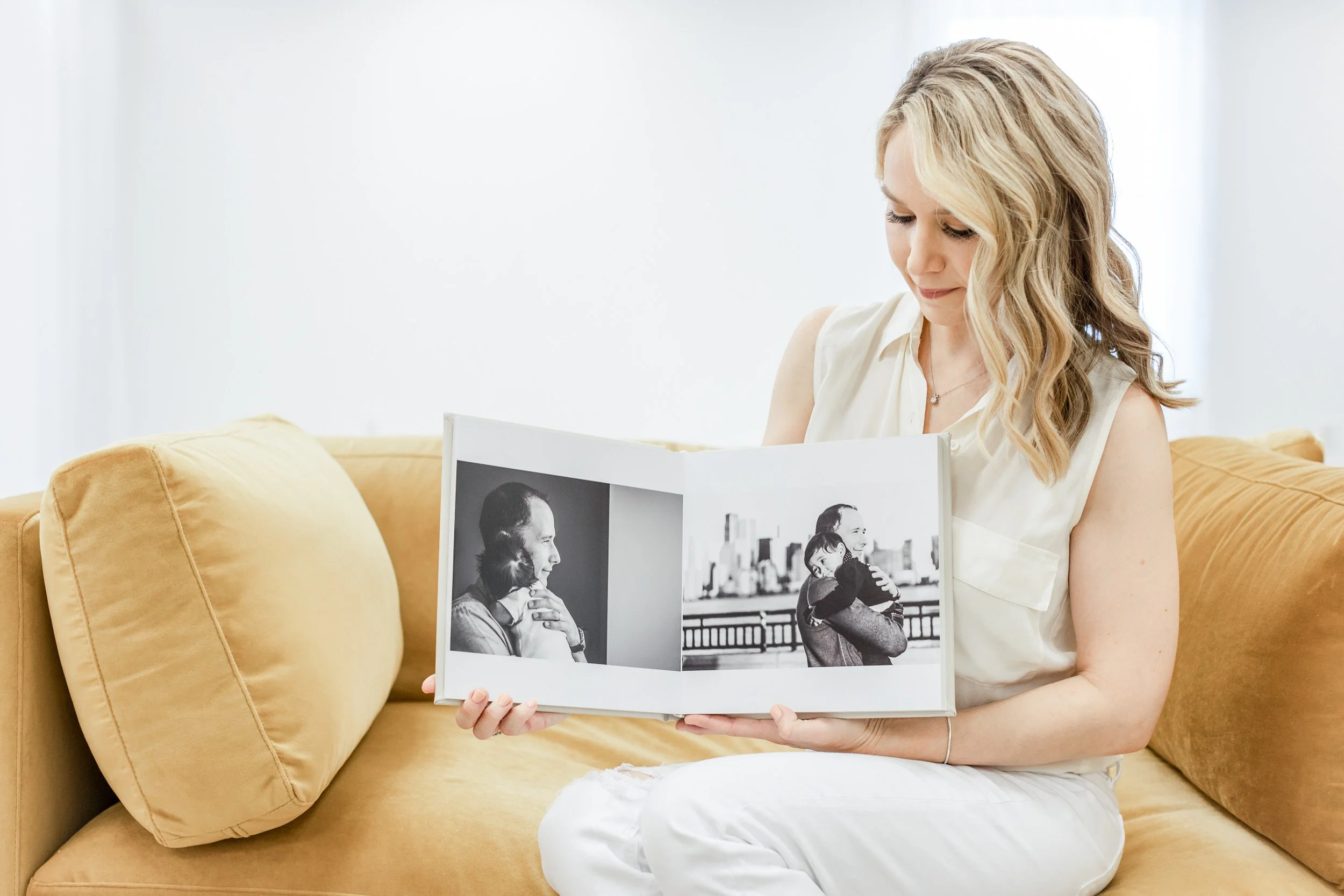 Newborn and family photographer with blonde hair sitting on a yellow sofa in luxe studio while looking at a photo album with black and white photos of a man hugging his newborn son.