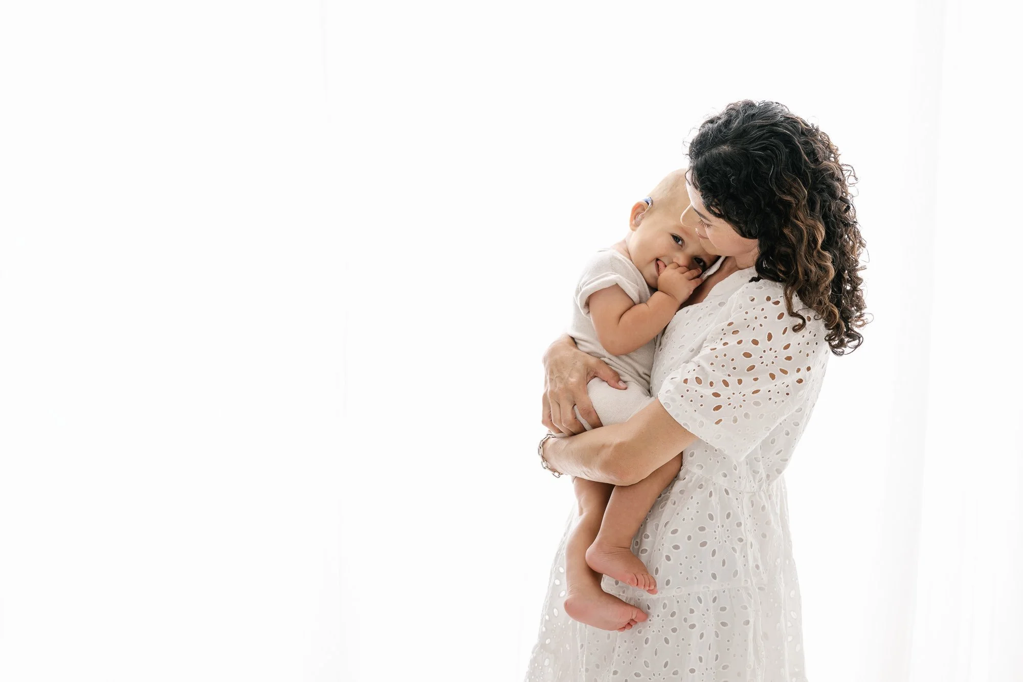 Mother in white eyelet dress cuddling baby for first birthday portraits in bright white NJ studio