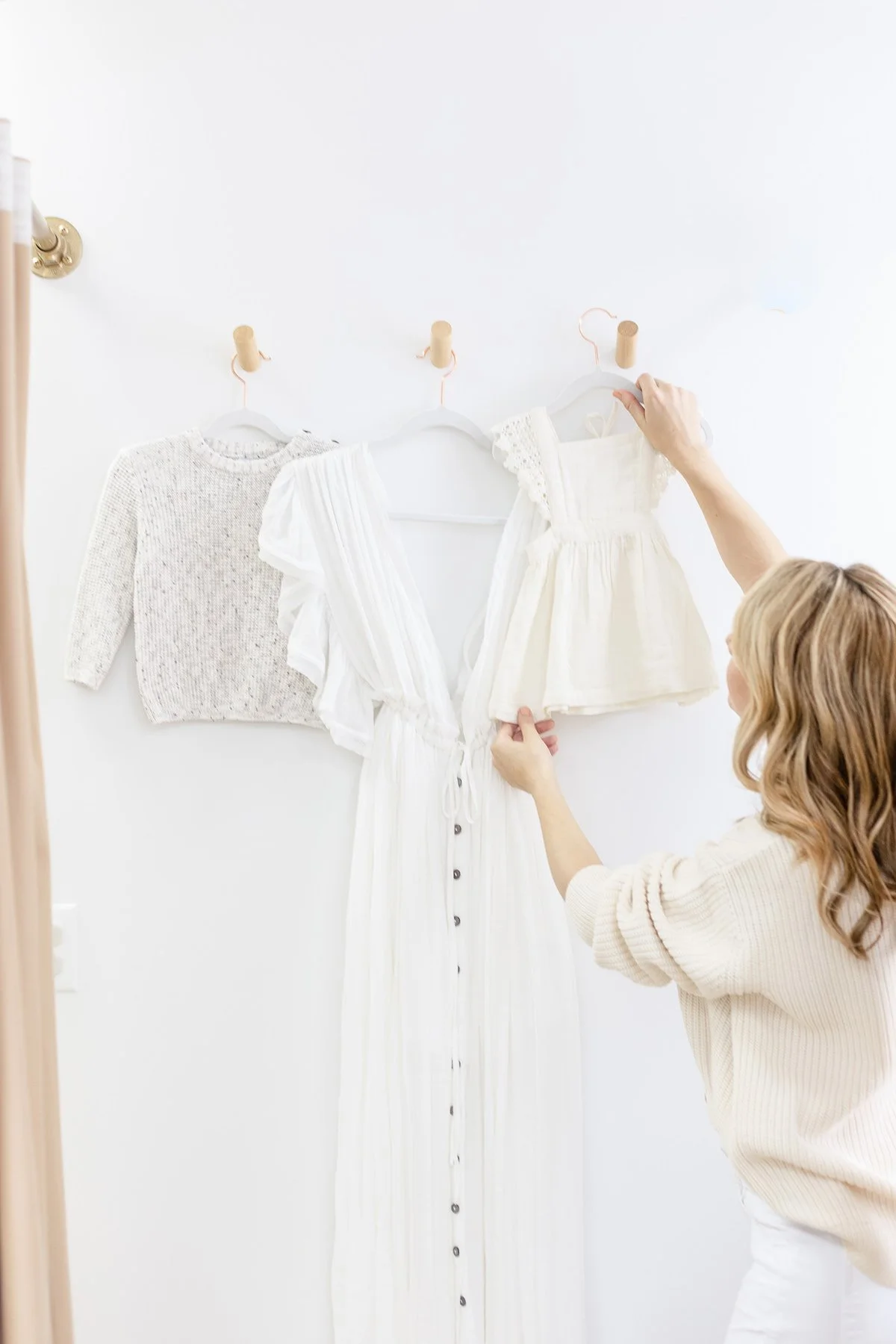 NJ family photographer Nicole Hawkins wearing cream knit sweater arranging two white dresses in dressing room for photo clients.