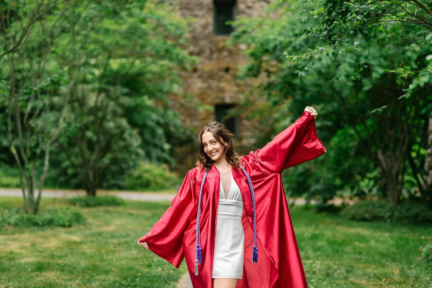 Senior wearing white dress, smiling, and celebrating in her red graduation gown at Cross Estate Gardens